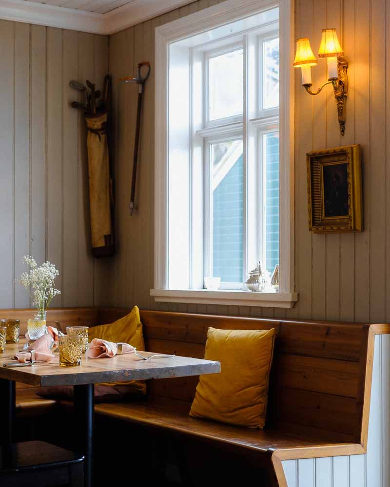 Coin repas avec une fenêtre à l'arrière, un banc en bois avec des coussins jaunes, une table en bois avec des verres, un vase avec des fleurs, une lampe murale et une décoration suspendue.