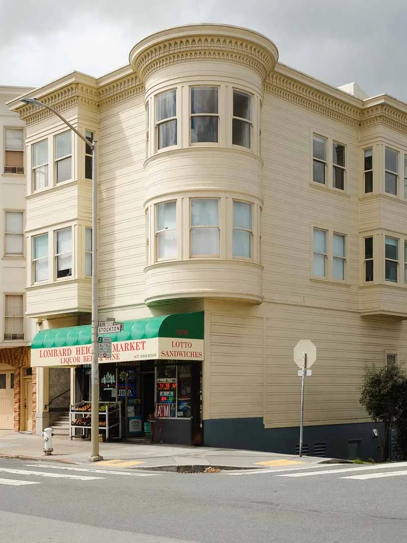 Une épicerie située en bas d'un bâtiment résidentiel de style victorien avec des fenêtres en baie, un auvent vert et une enseigne indiquant Lom bard Height Market et Lotto Sandwiches.