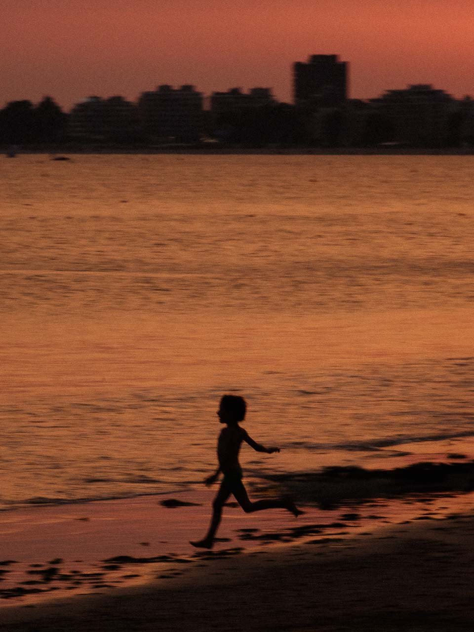 Un enfant court sur la plage au coucher du soleil, avec un ciel orange et des silhouettes de bâtiments de la baule à l'horizon.
