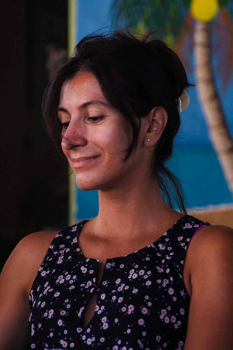 Une femme avec des cheveux noirs, portant un haut à fleurs, qui sourit doucement, fond coloré avec des éléments naturels.