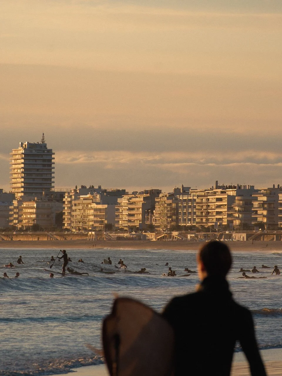 Une plage au coucher du soleil avec de nombreuses personnes pratiquant le surf ou nageant, et en arrière-plan, un skyline avec des bâtiments modernes.
