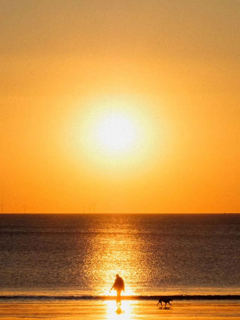 Une personne et un chien marchant sur la plage au coucher du soleil, avec un ciel orange et le soleil brillant au-dessus de l'horizon