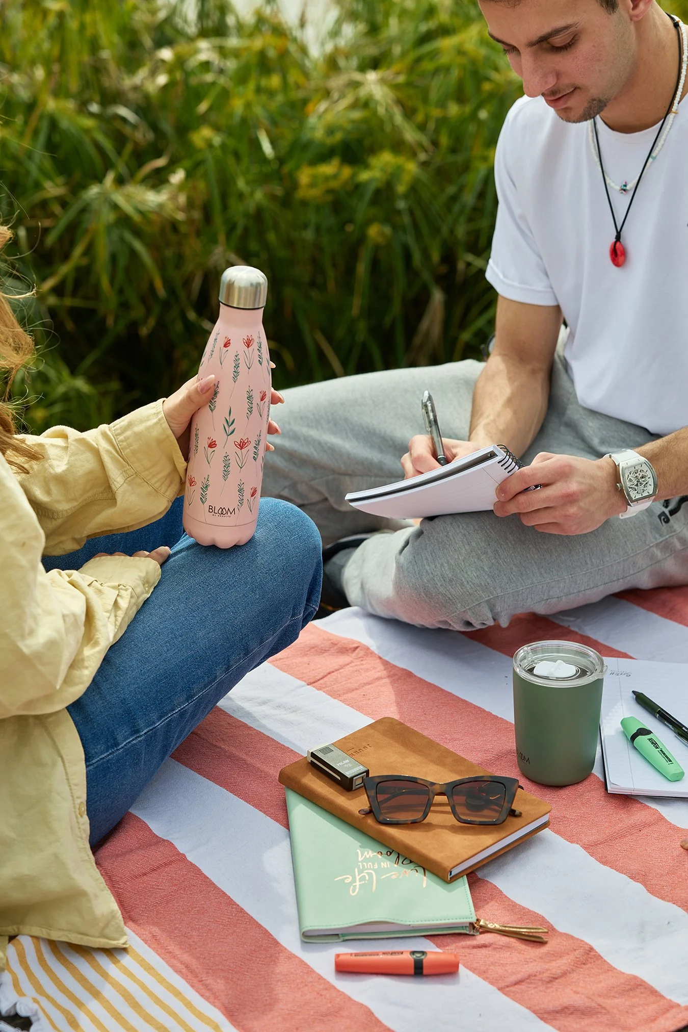 Two people sitting on a blanket outdoors, one holding a pink water bottle with a floral pattern, and the other taking notes in a small notebook. The blanket has a striped pattern, and there are various items around, including sunglasses, a green cand