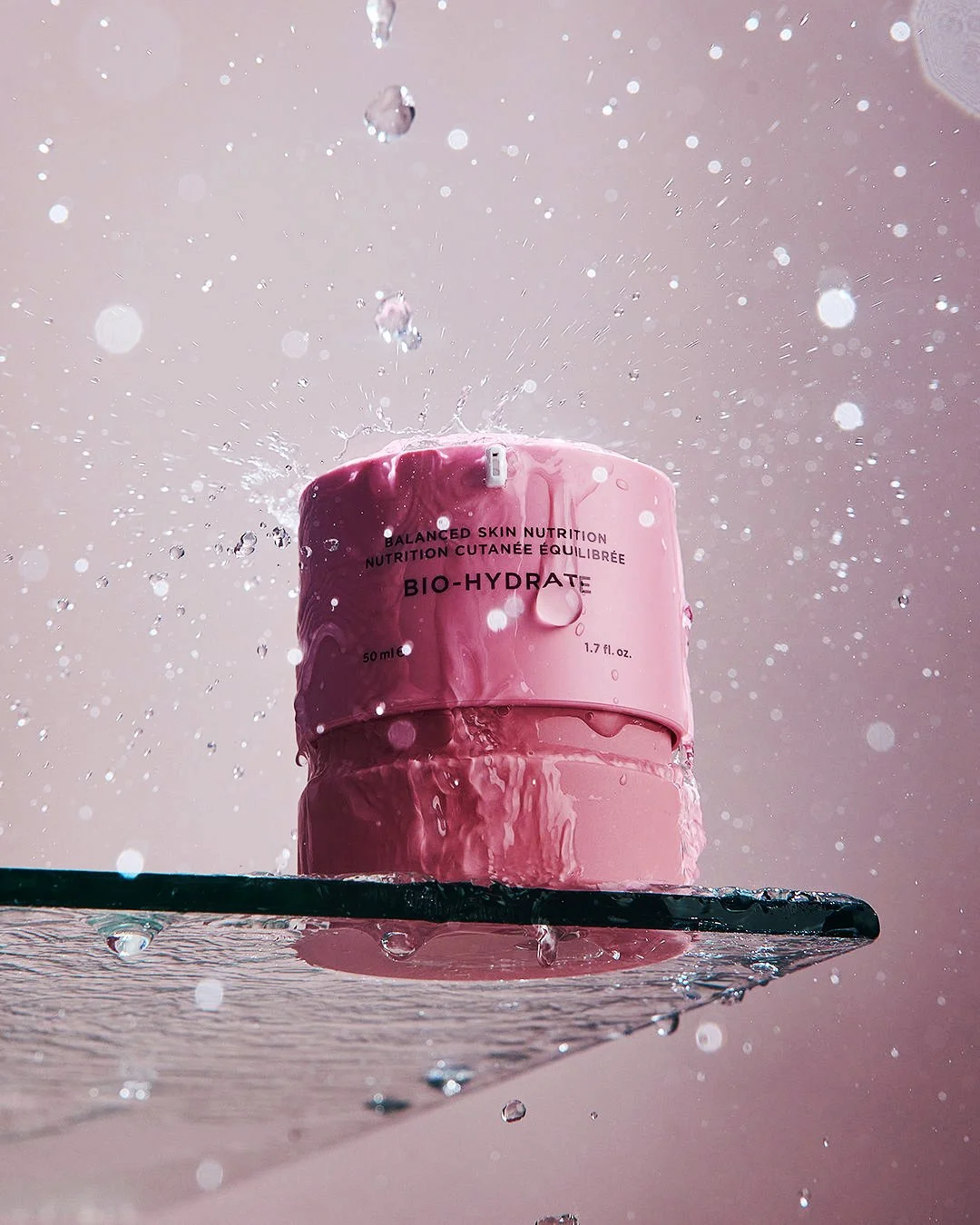 Pink skincare cream container labeled 'Bio-Hydrate' with water splashing on it, placed on a black surface with water droplets around, against a light pink background.