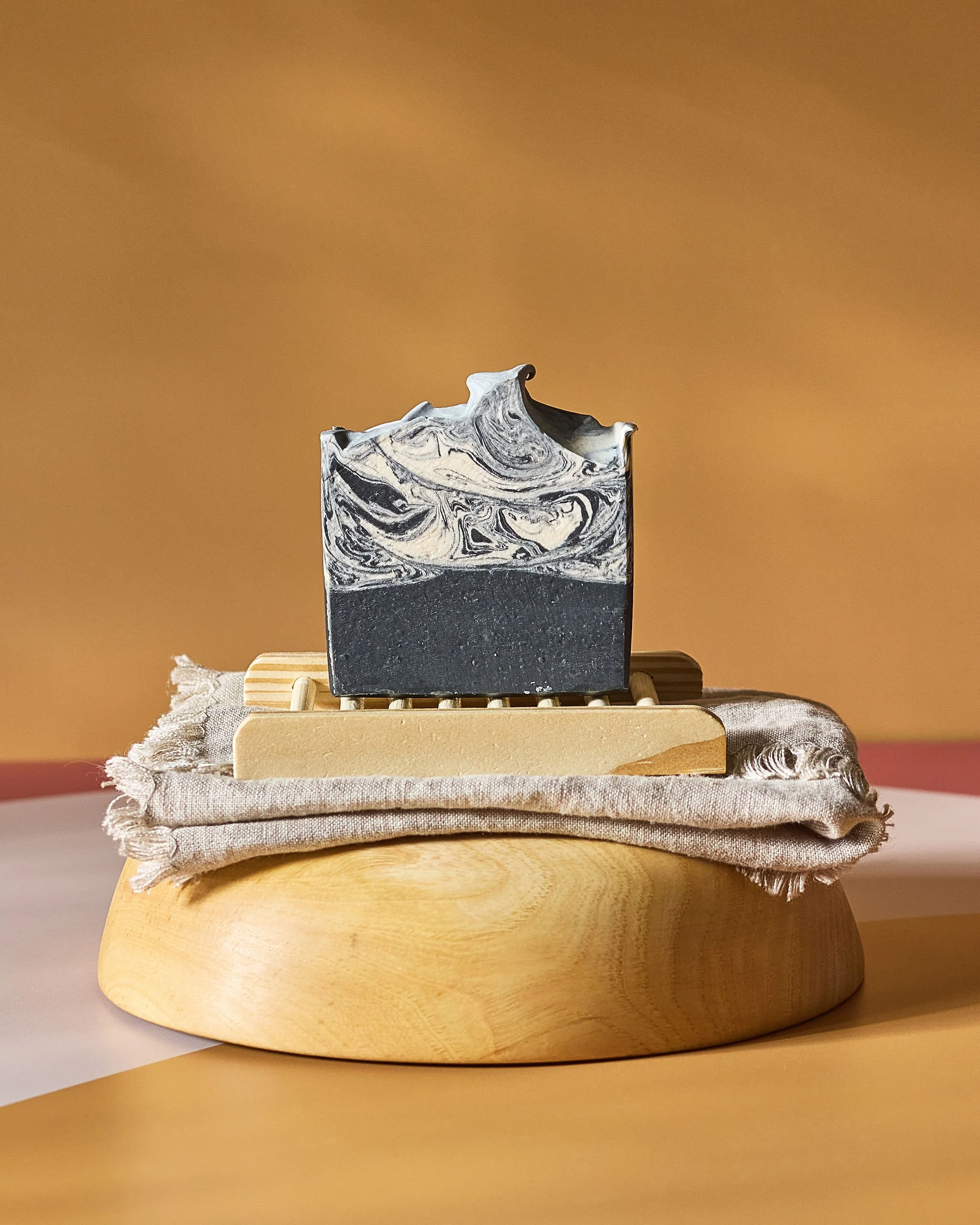 A black and white marbled soap atop a wooden soap dish, placed on a beige cloth on a rounded wooden base.