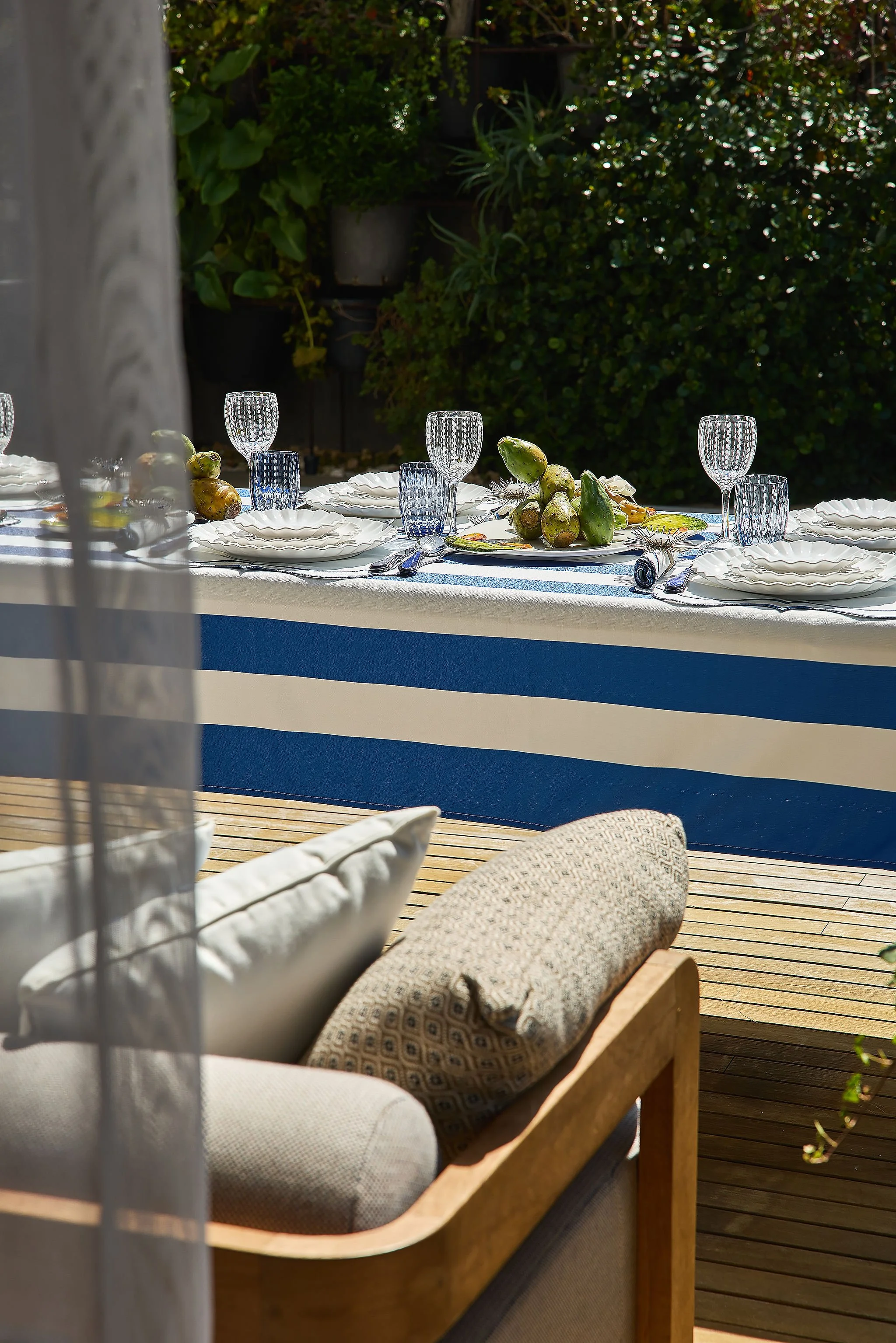 An outdoor dining table set with plates, glasses, and a centerpiece of artichokes, with greenery in the background and a wooden deck surface.