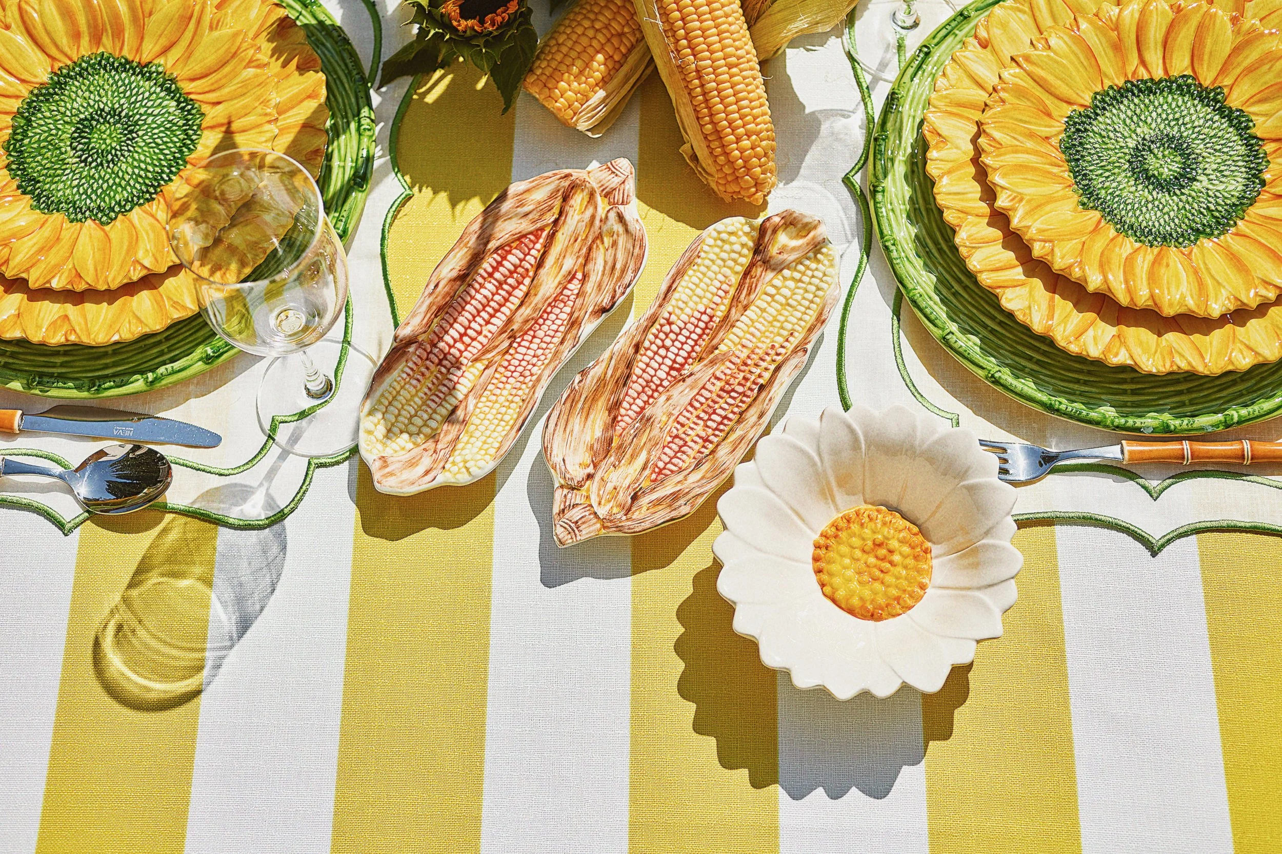 A summer outdoor table setting with yellow sunflower-shaped plates, a white flower-shaped bowl, ears of cooked corn on the cob, and a glass of white wine on a yellow and white striped tablecloth.
