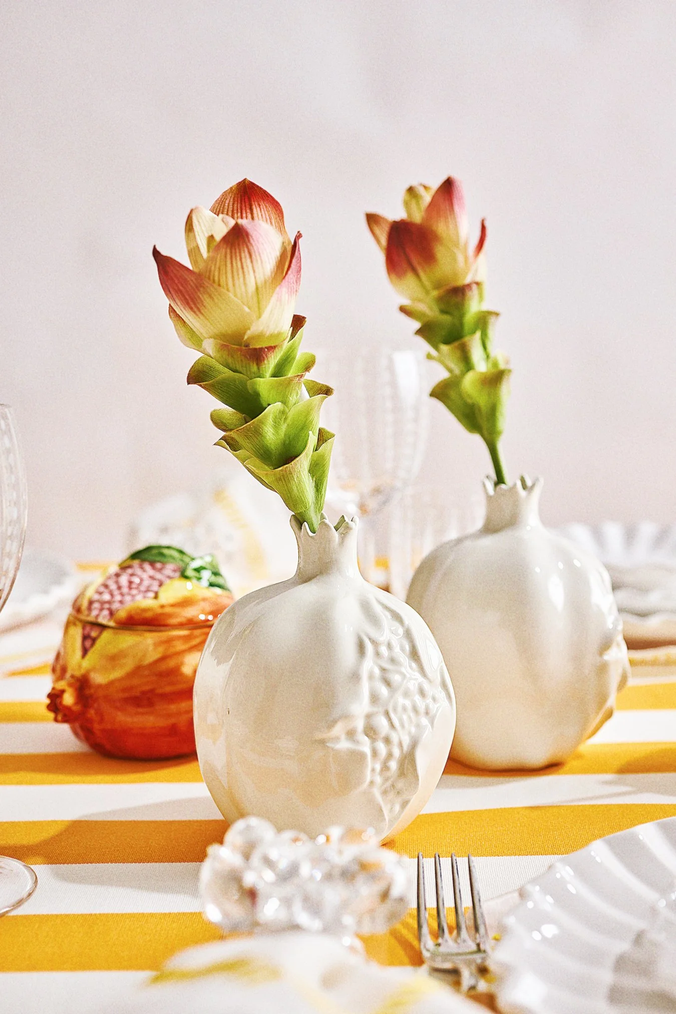 Two white ceramic vases with floral patterns holding pink and green flowers on a yellow and white striped tablecloth.