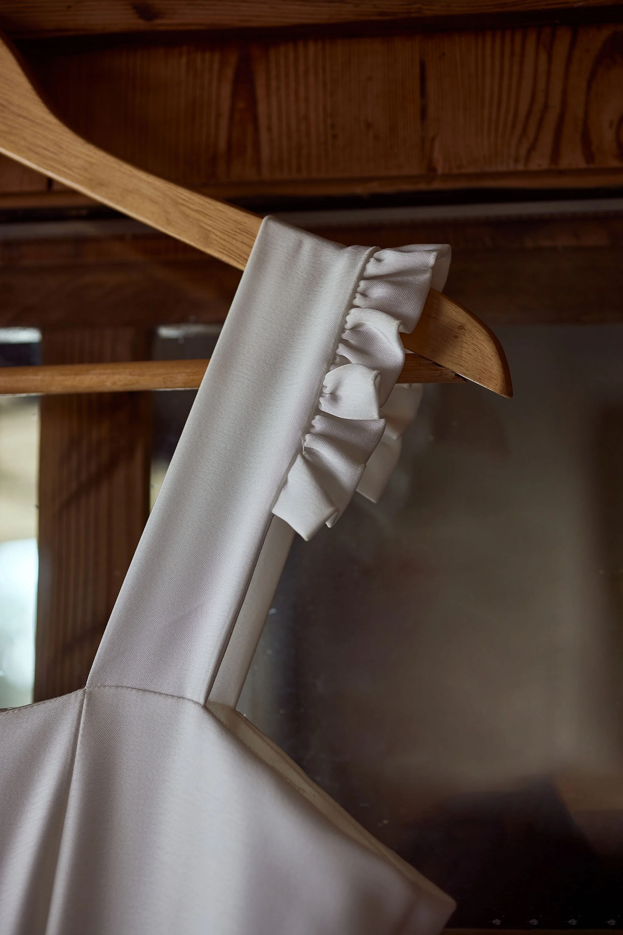 Close-up of a white satin wedding or special occasion dress with a ruffled strap, hanging from a wooden chair.