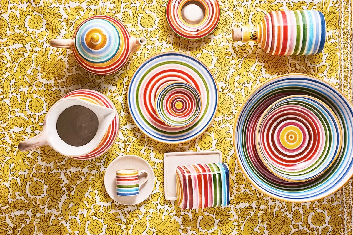 Colorful striped dinnerware, teapot, mug, and plates on a yellow floral tablecloth.