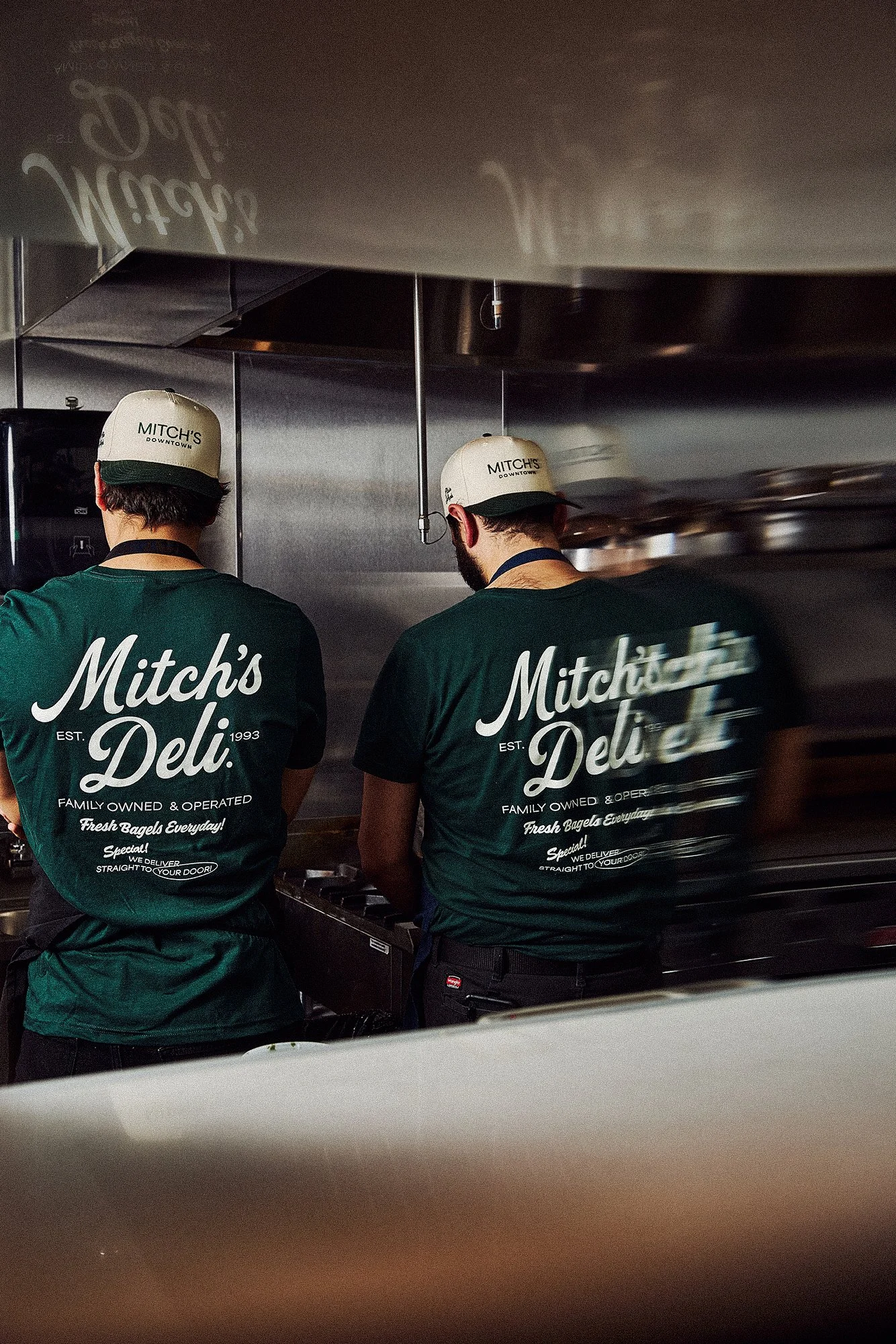 Two employees working behind the counter at Mitch's Deli, both wearing dark green t-shirts and beige hats with the deli's logo, in a kitchen setting with stainless steel appliances.