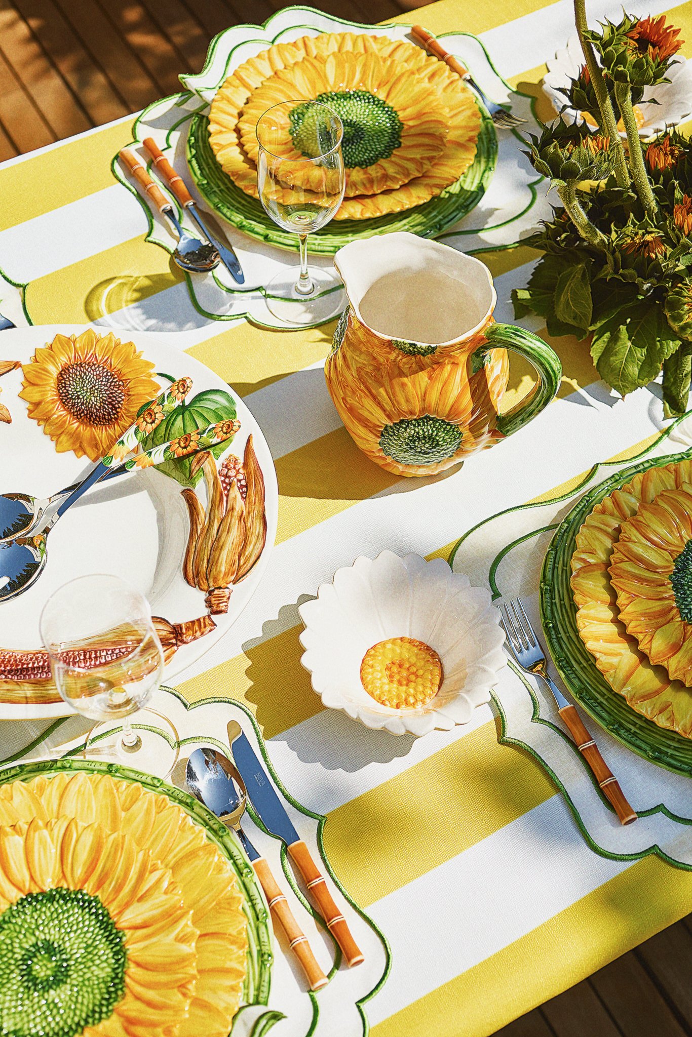 A table set with sunflower-themed dishes and accessories, including sunflower-patterned plates, a sunflower-painted pitcher, and matching placemats, along with clear wine glasses, silverware, and a floral centerpiece.