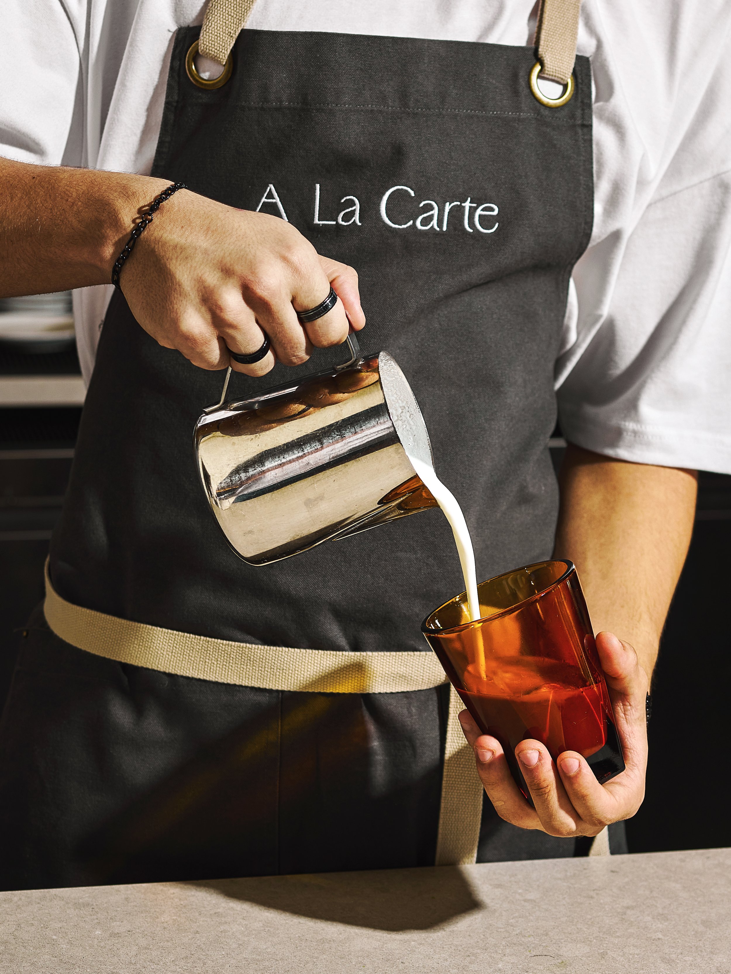 A person wearing an apron with 'A La Carte' embroidery pouring milk into a glass. cafe place in Miami Hallandale beach