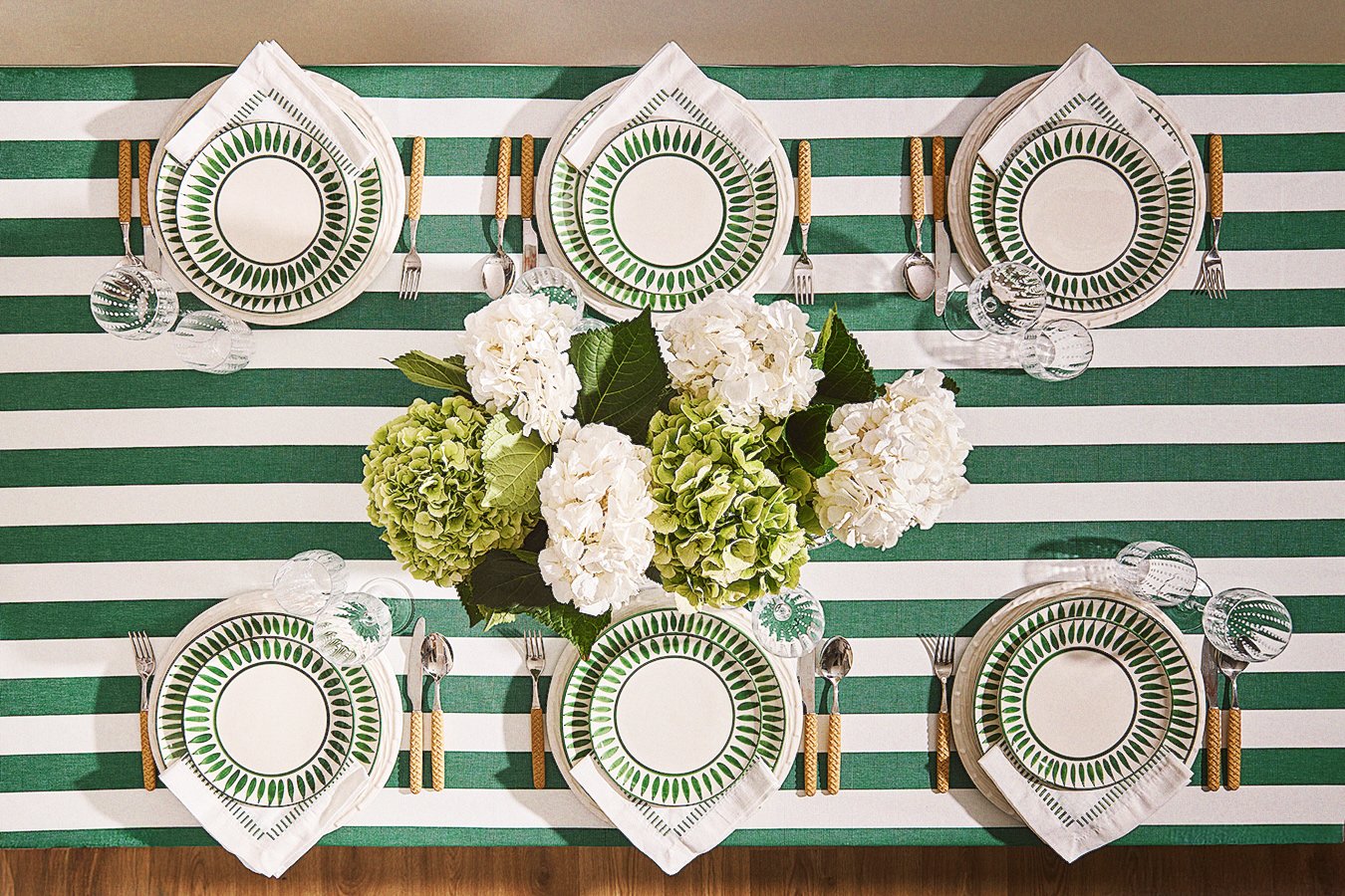 Table setting with six plates featuring green leaf patterns, silverware with beige handles, and clear glassware, with a centerpiece of white and green hydrangeas on a green and white striped tablecloth.