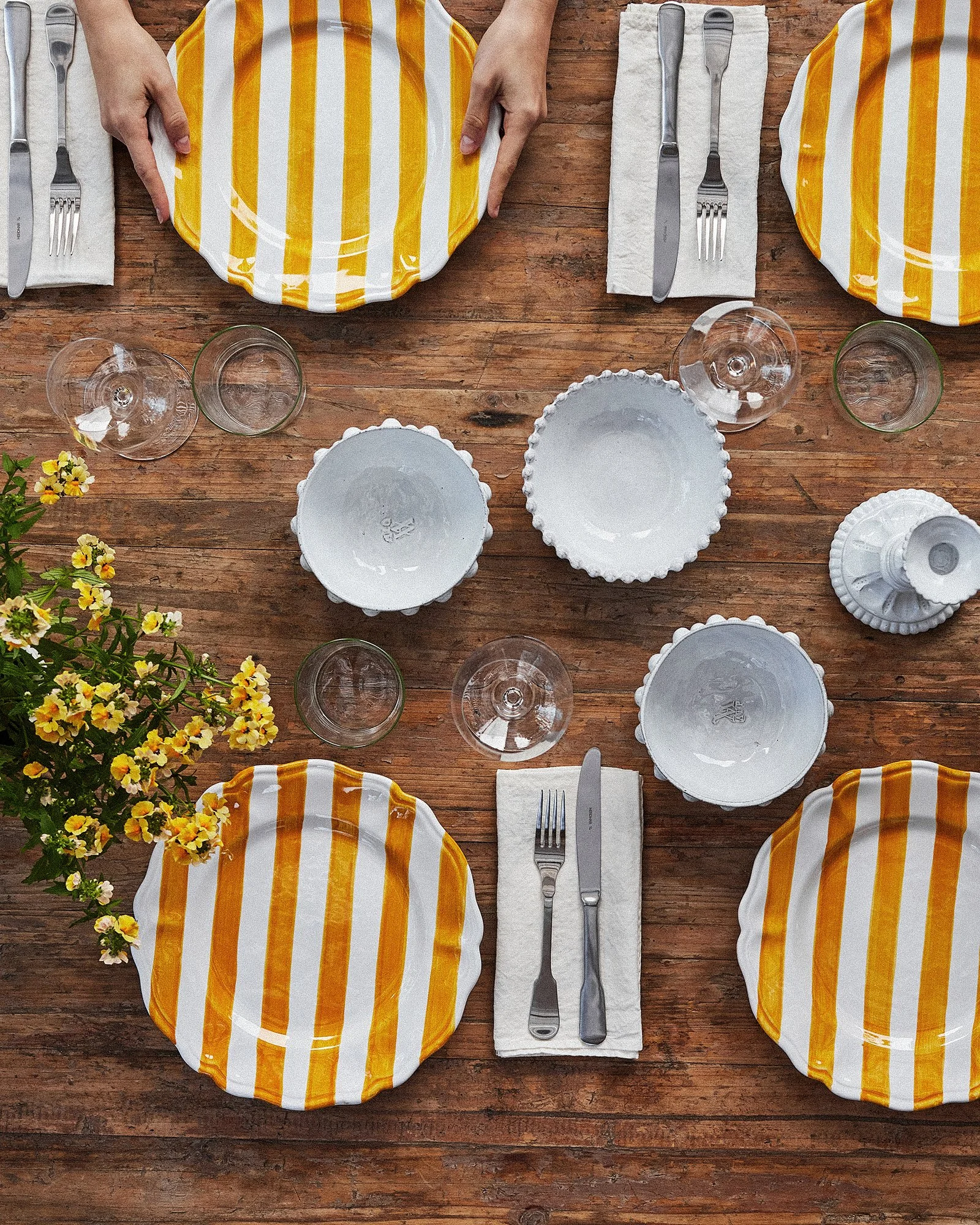 Top view of a wooden dining table set for four with yellow and white striped plates, clear wine glasses, white bowls, silverware on white napkins, and a flower arrangement.