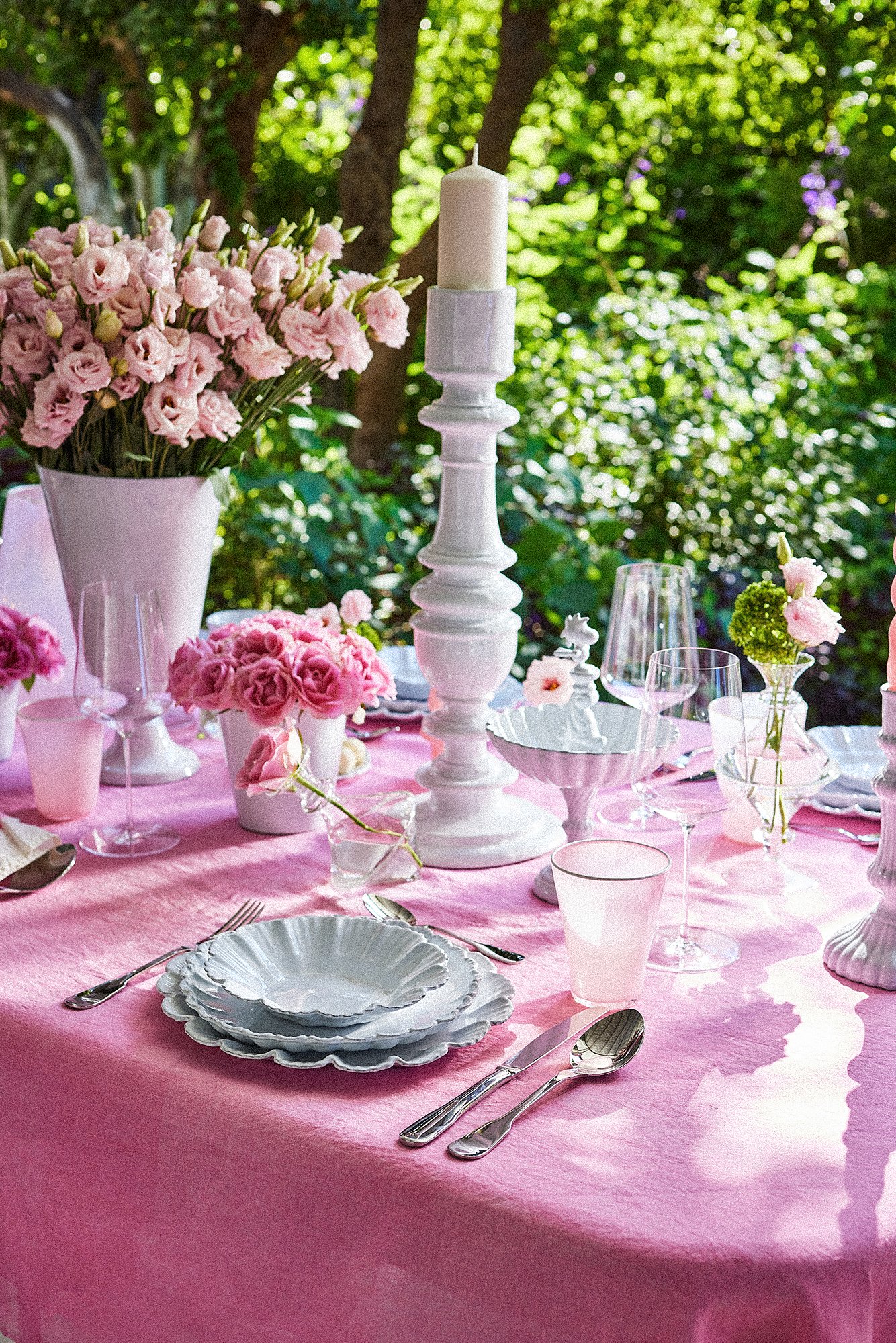 A table set outdoors with pink tablecloth, floral arrangements, white plates, silverware, wine glasses, and a white candle in a decorative holder, surrounded by lush greenery.