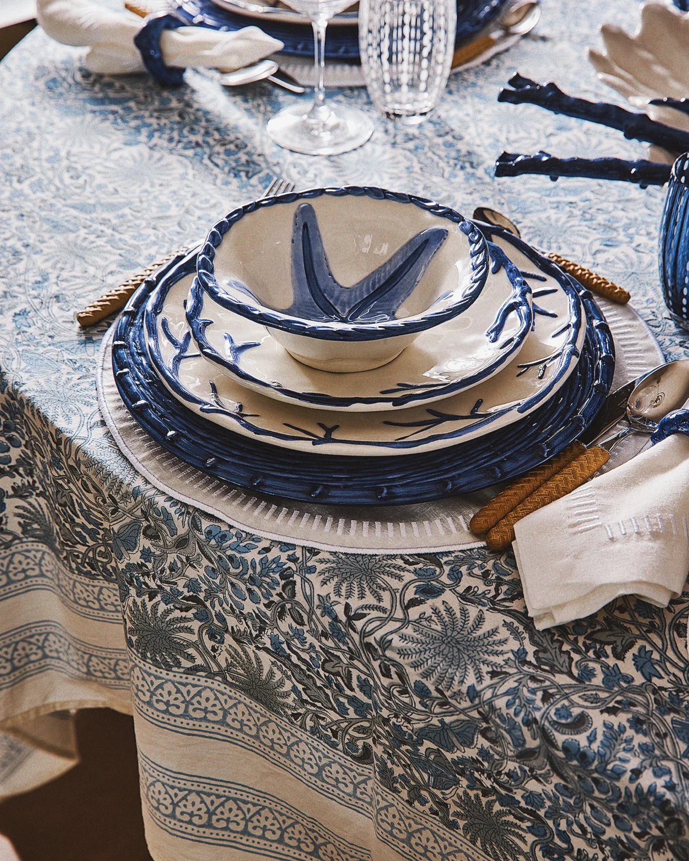 A table setting with blue and white patterned ceramic plates and bowls, a glass of water, a wine glass, and silverware on a tablecloth with matching pattern.