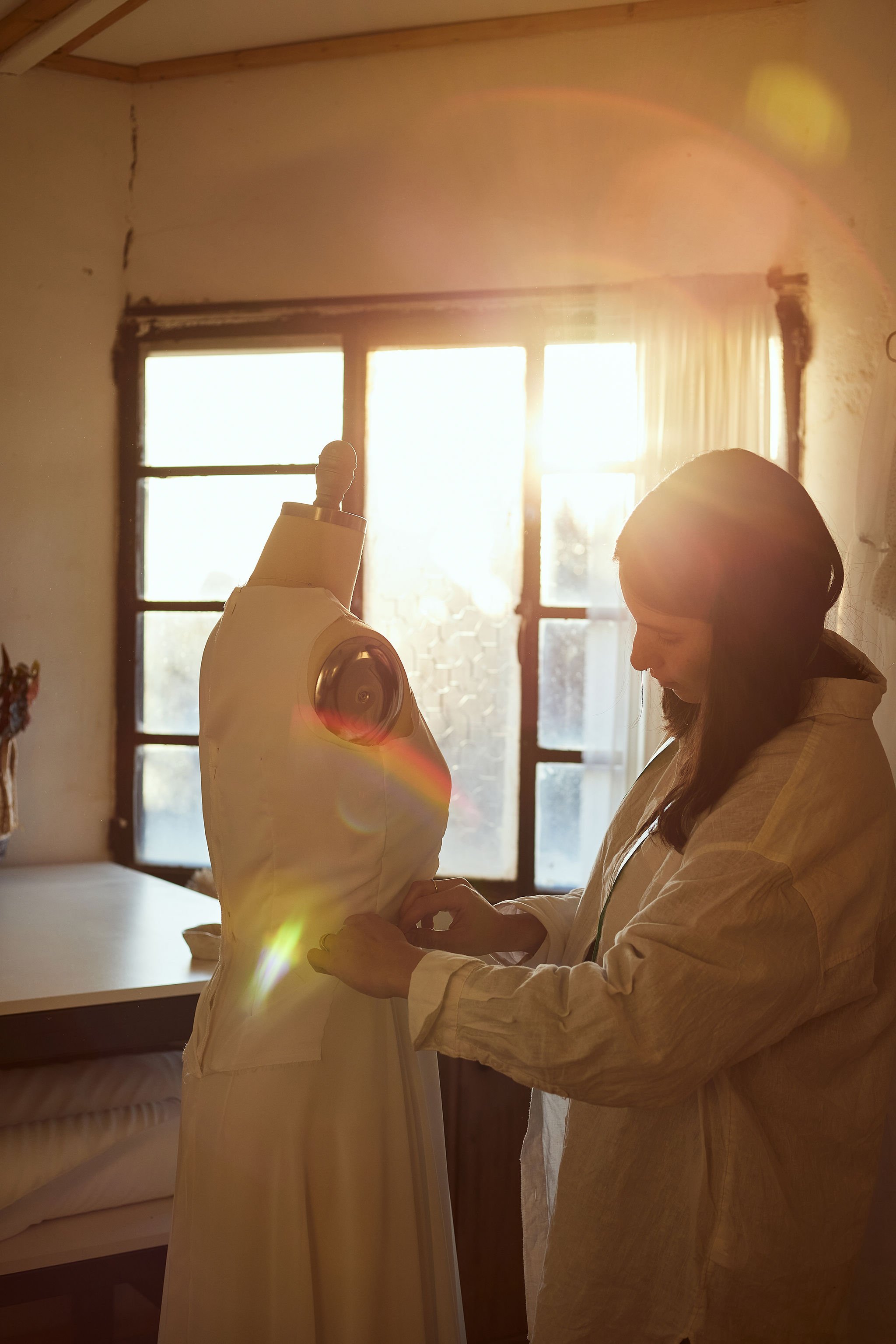 A woman working on a dress mannequin indoors with sunlight coming through a large window in the background.