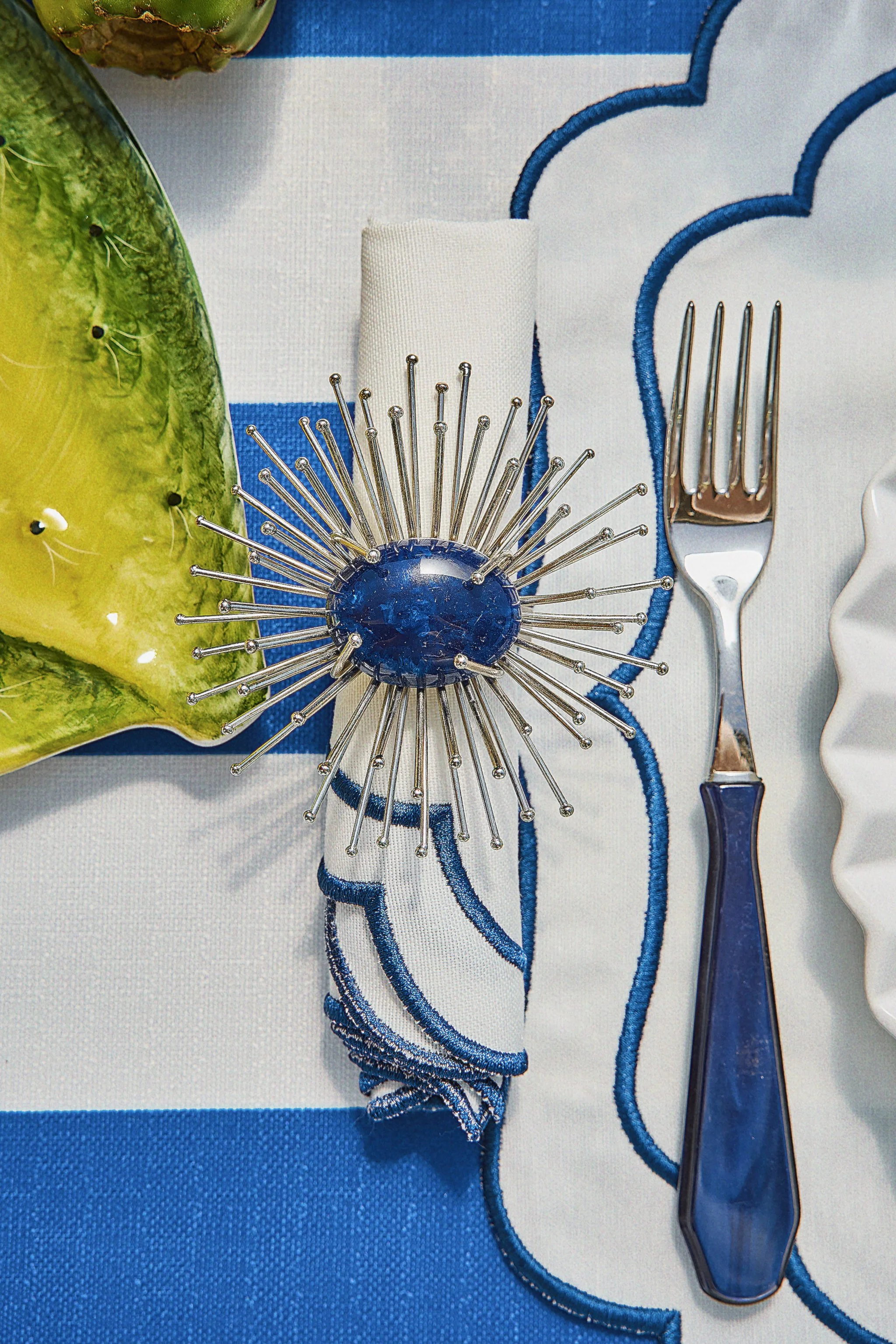 A table setting with a decorative silver and blue starburst salt shaker, a blue and white embroidered napkin, a silver fork with a blue handle, and a yellow-green glazed ceramic plate, on a white and blue tablecloth.