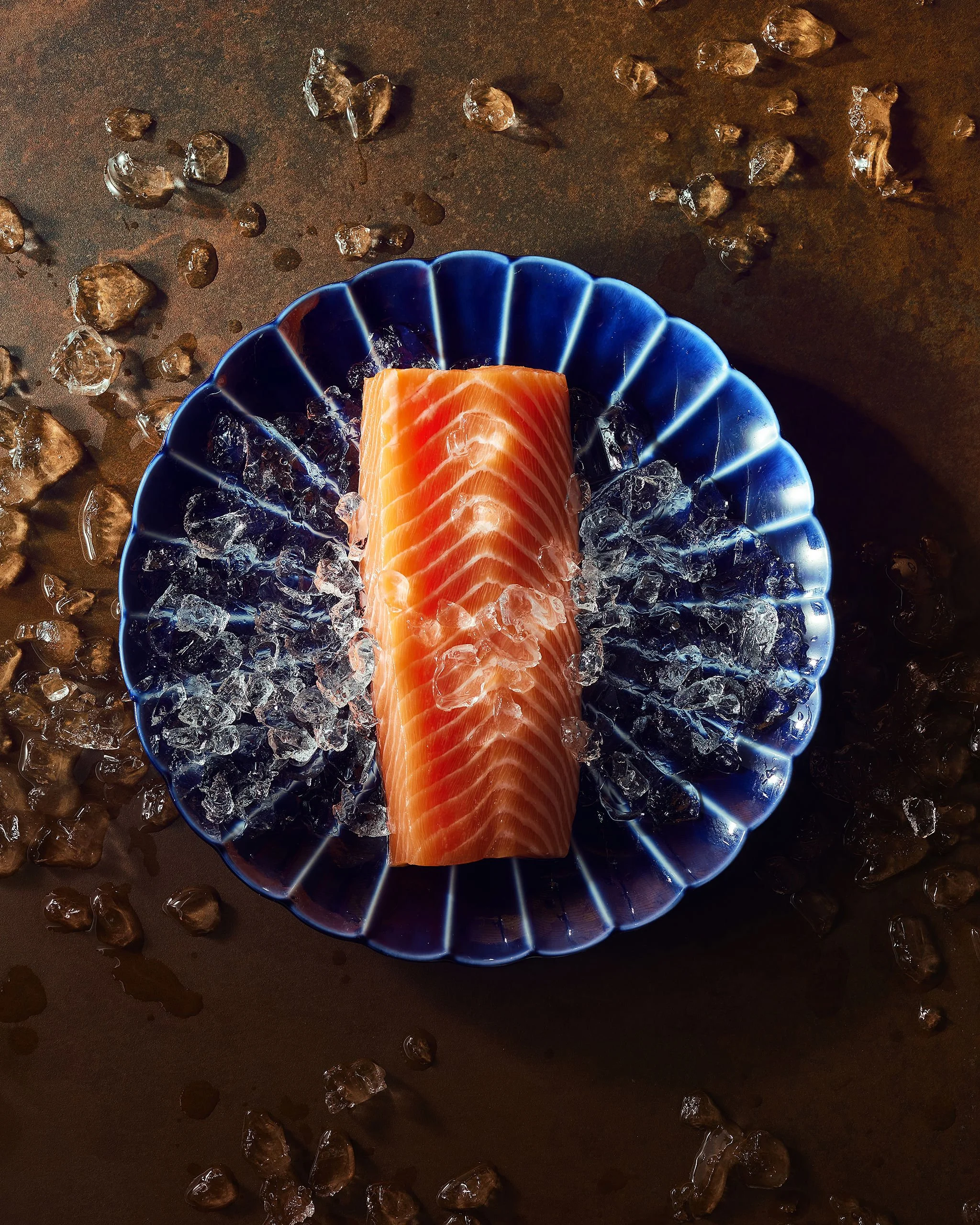 Sliced salmon fillet on a blue glass dish filled with ice, surrounded by scattered ice cubes, on a dark brown surface.