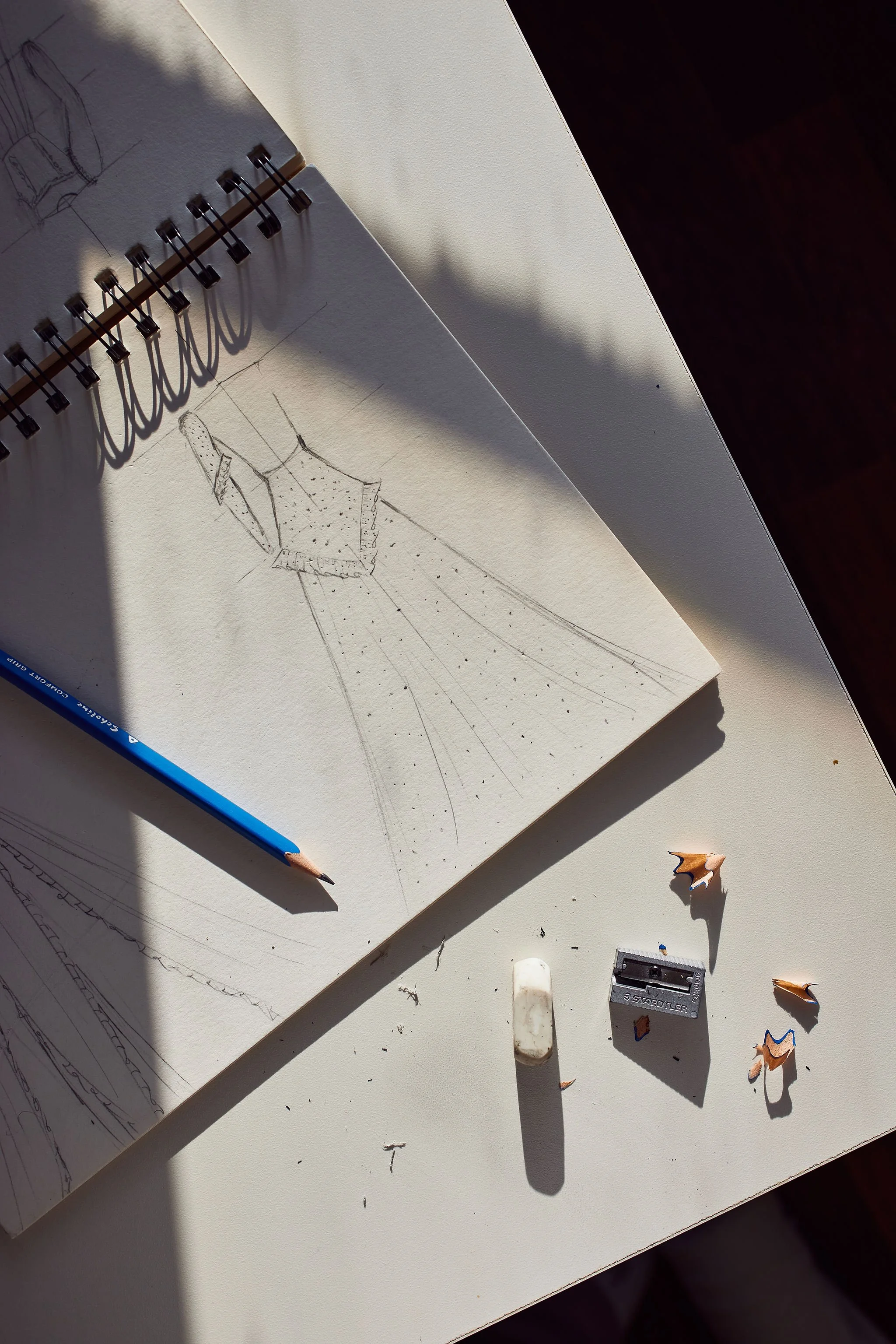 Sketchbook with fashion design sketches, blue colored pencil, eraser, pencil sharpener, and shavings on a white desk illuminated by sunlight.