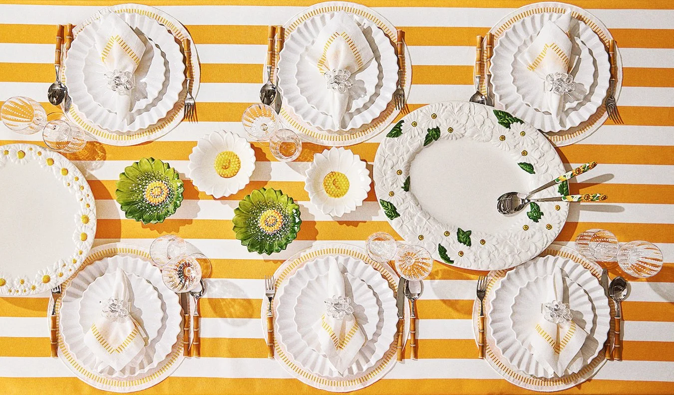Table setting with white and gold plates, napkins, and cutlery, decorated with small glass candles and floral centerpieces on a yellow and white striped tablecloth.