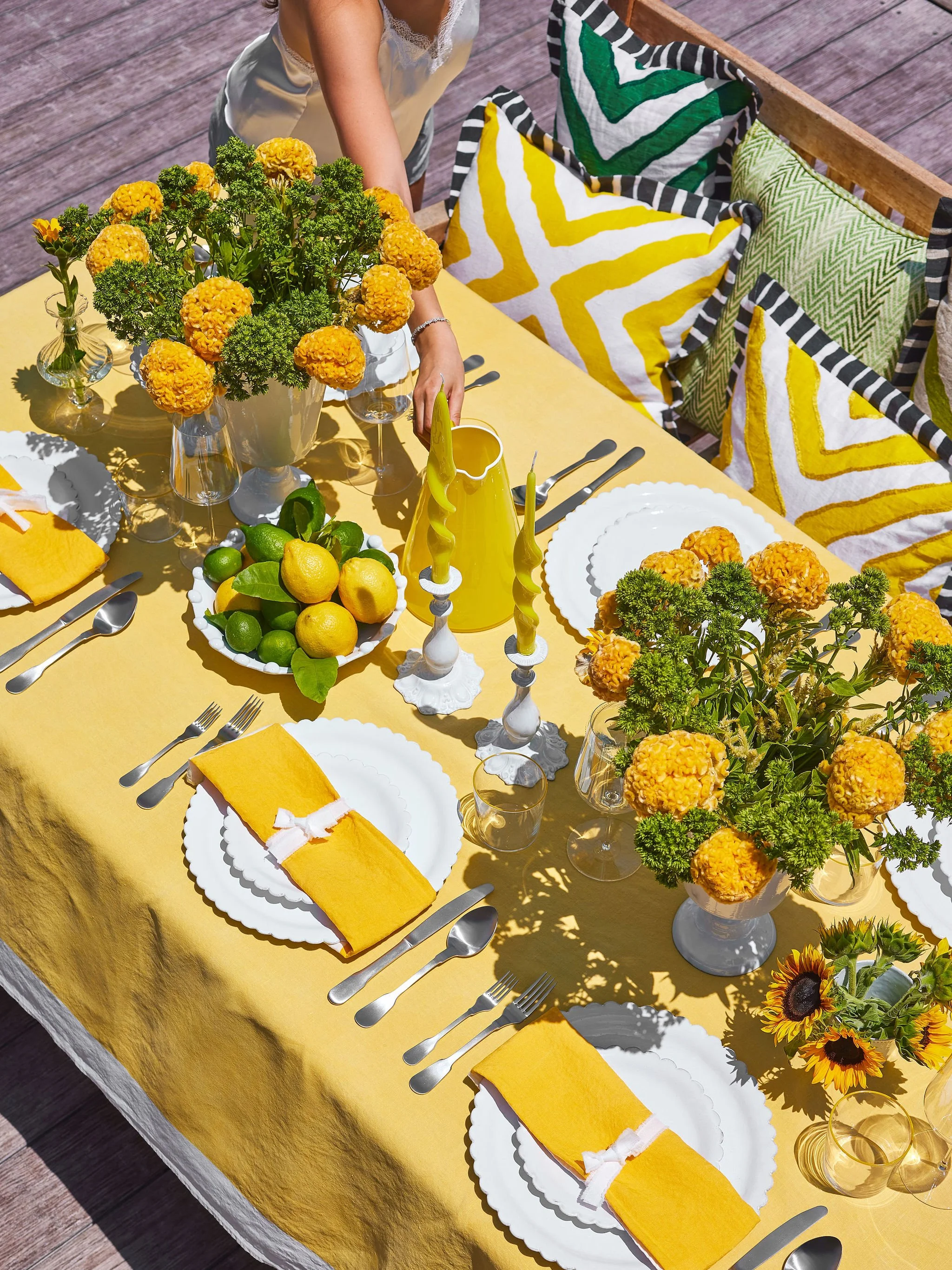 A brightly decorated outdoor dining table with a yellow tablecloth, yellow napkins, white plates, silverware, and glasses. The table has yellow and orange floral centerpieces, a bowl of lemons and limes, and yellow candles. A person is reaching for s