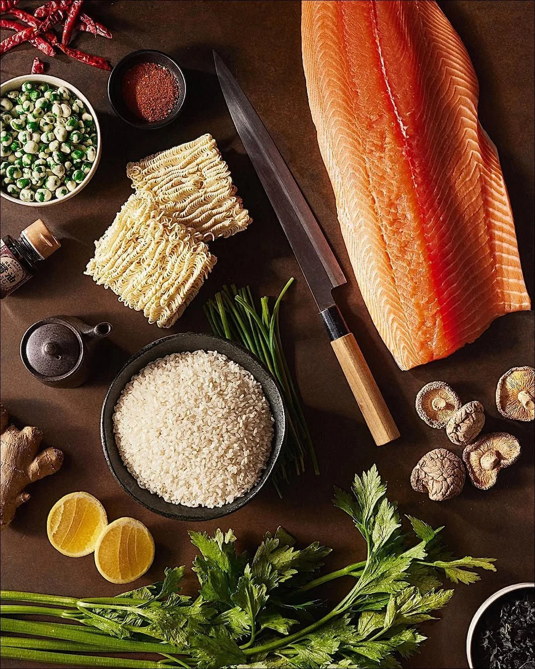Fresh ingredients for cooking, including a large piece of salmon, rice, instant ramen noodles, green onions, ginger, lemon slices, shiitake mushrooms, dried red chili peppers, black sesame seeds, and various seasonings, arranged on a dark surface wit