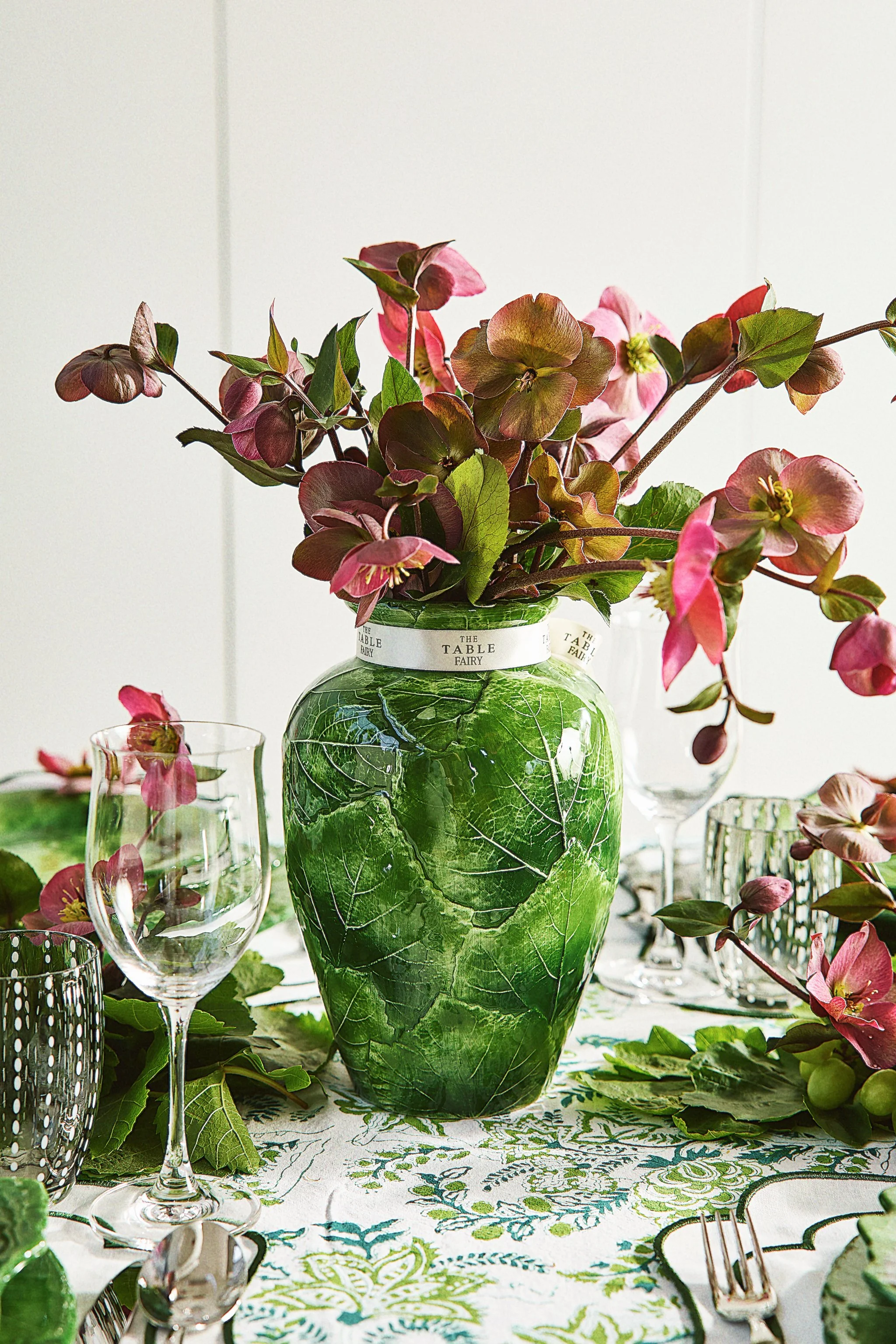 Decorative table setting with a green leafy vase filled with pink and purple flowers, surrounded by glassware, silverware, and a green leaf-patterned tablecloth.