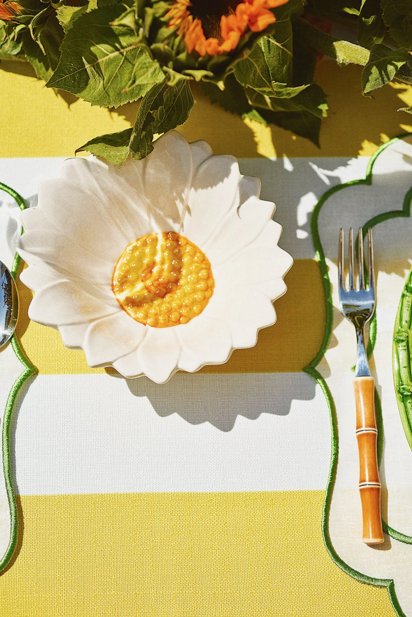 A white ceramic dish shaped like a flower with a yellow jam in the center, placed on a yellow and white tablecloth with green decorative placemats. A fork with a bamboo-style handle is next to the dish.