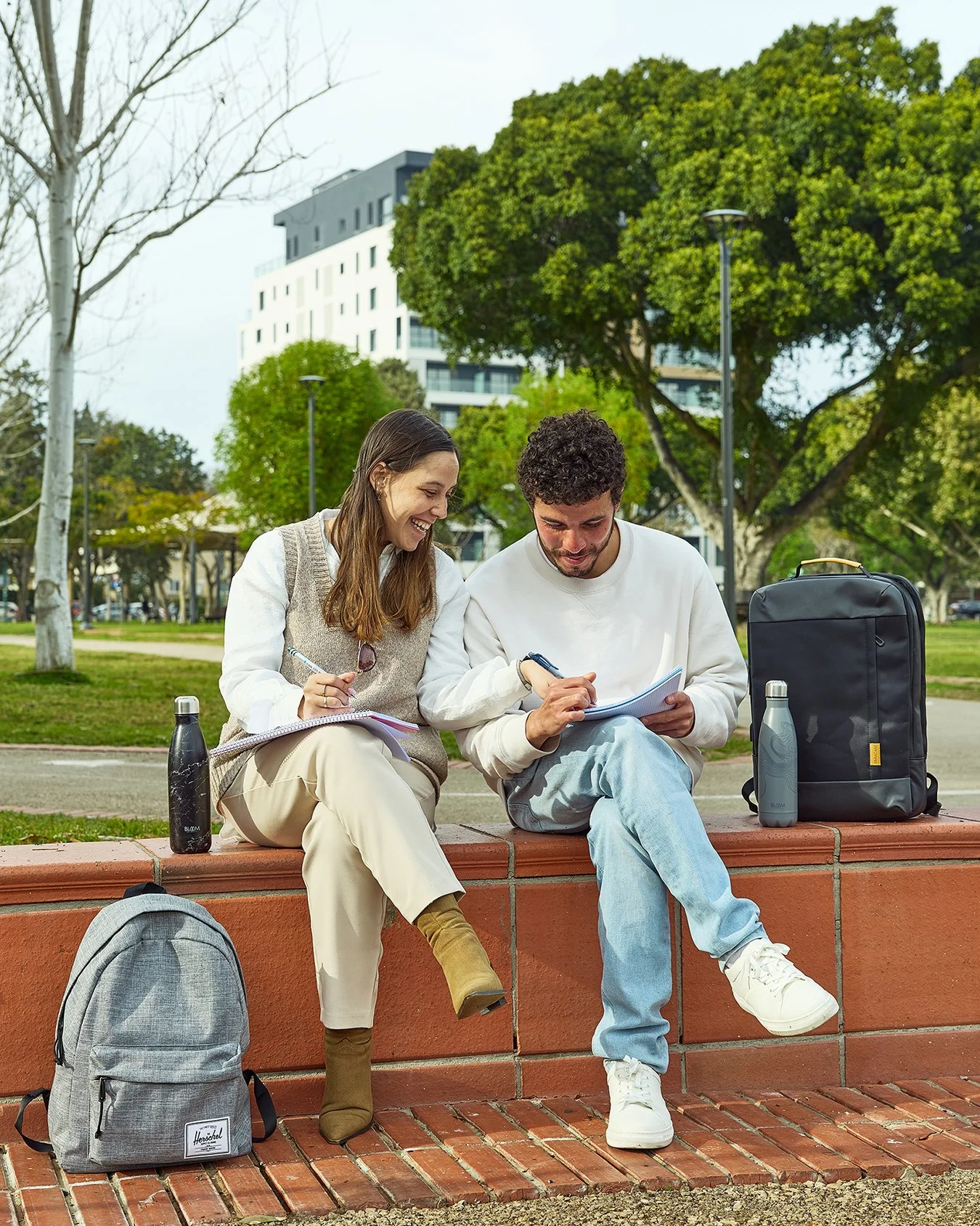 Two young adults, a woman and a man, sitting on a brick ledge in a park, smiling and working on notebooks together, with backpacks and water bottles nearby, trees and a building in the background.
