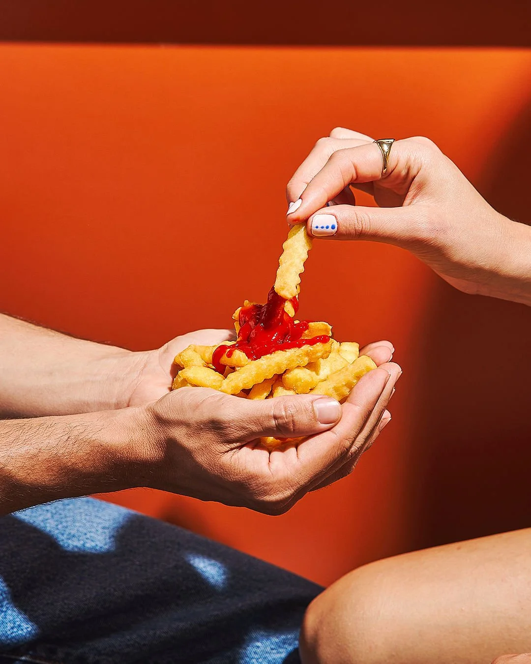 Close-up of two people sharing a plate of crinkle-cut French fries with ketchup, one person dipping fries into ketchup while the other holds the plate, with an orange background.