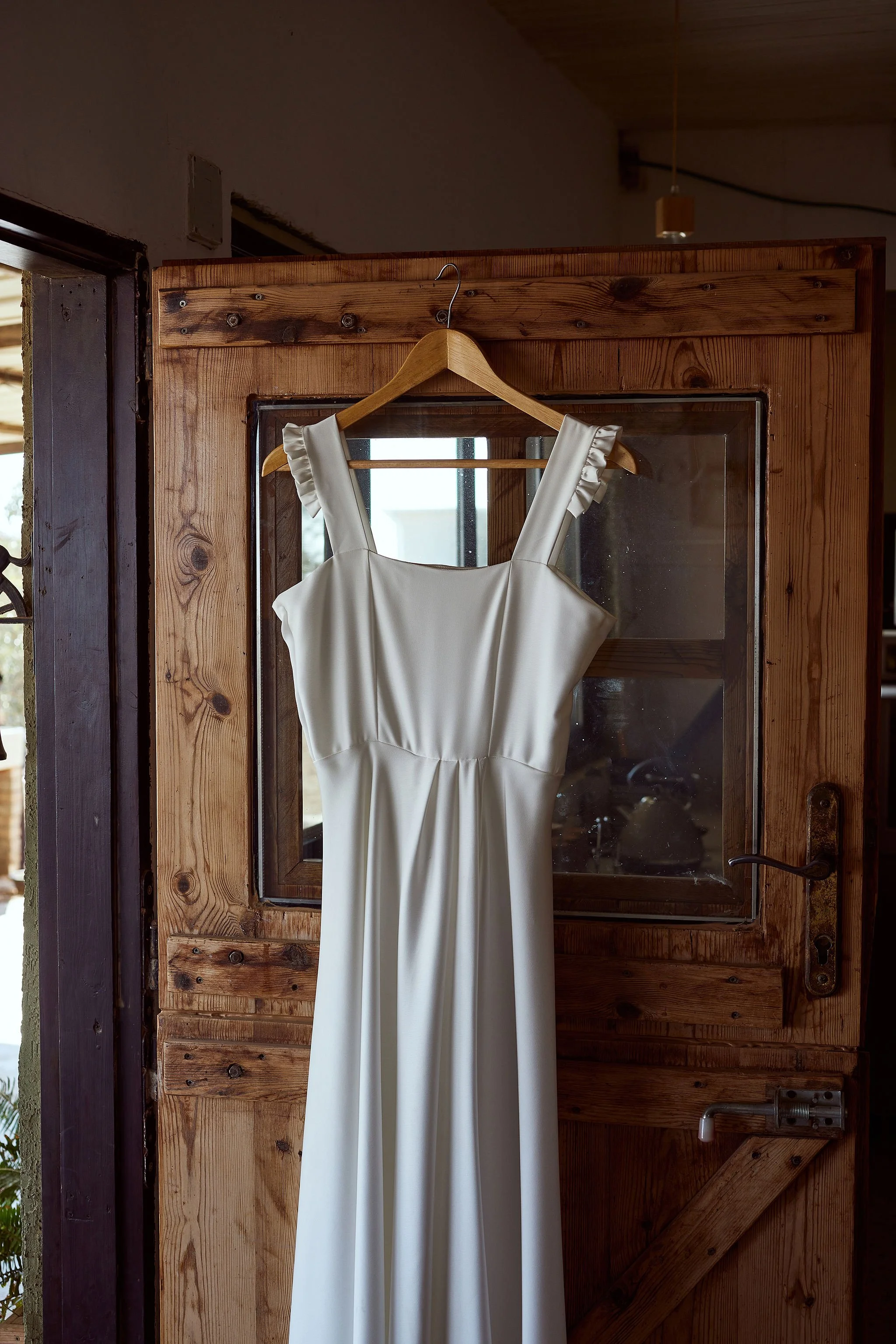 A white wedding dress hanging on a wooden hanger from a glass-paned rustic wooden door.