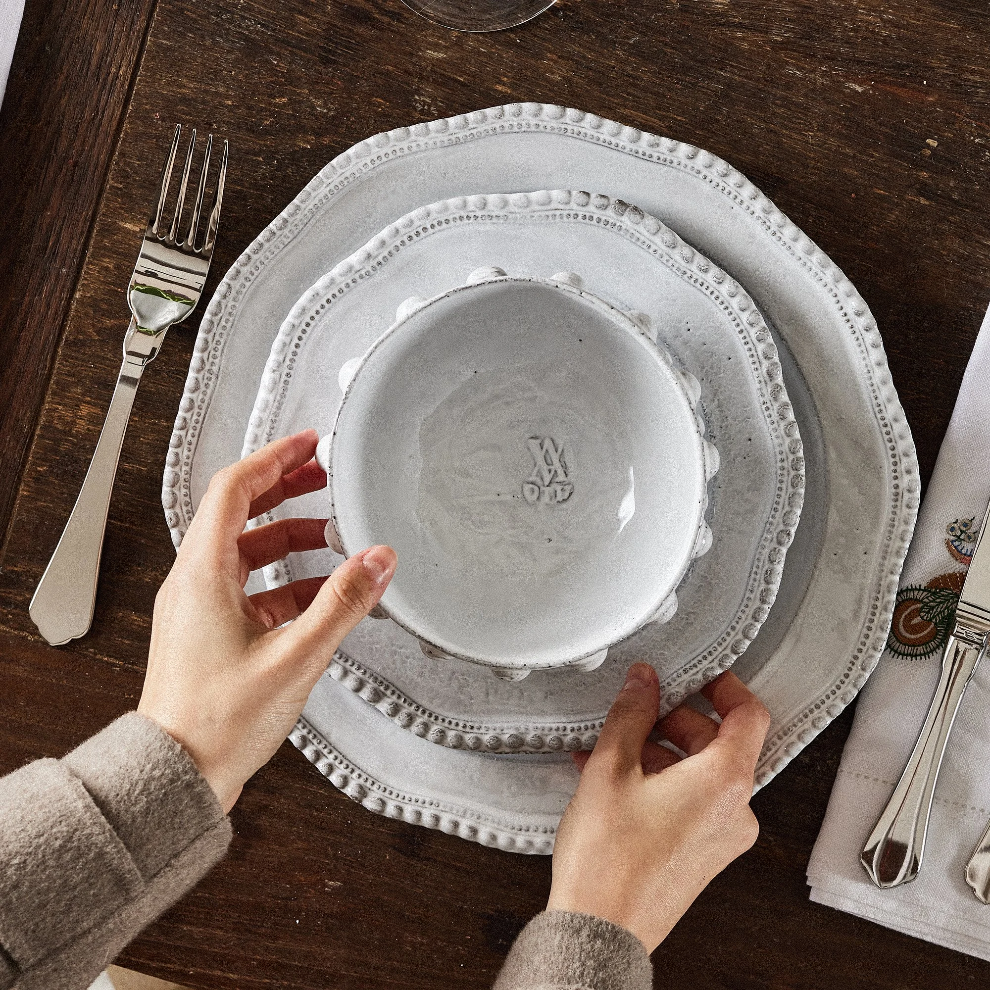 Hand placing a white ceramic bowl on a dinner plate at a table setting with a fork on the left and a knife on the right, on a dark wooden table with a white napkin.