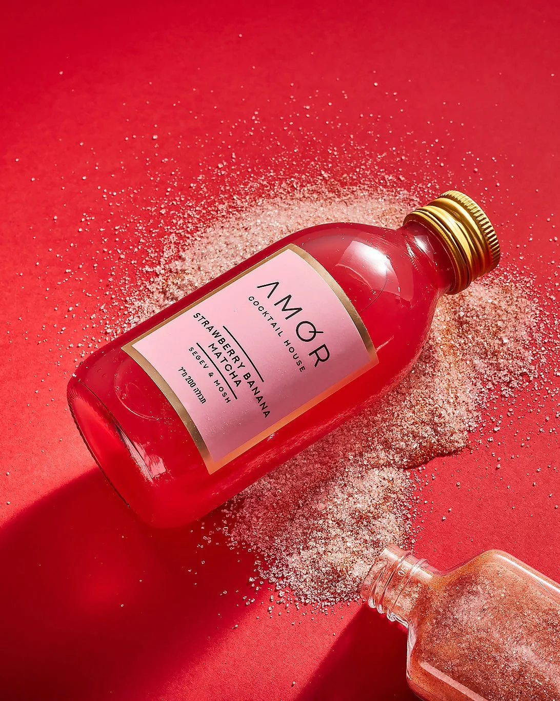 A pink bottle labeled 'AMOR Cocktail House' lying on a red surface, with pink sugar and a small glass bottle nearby, surrounded by pink sugar crystals.