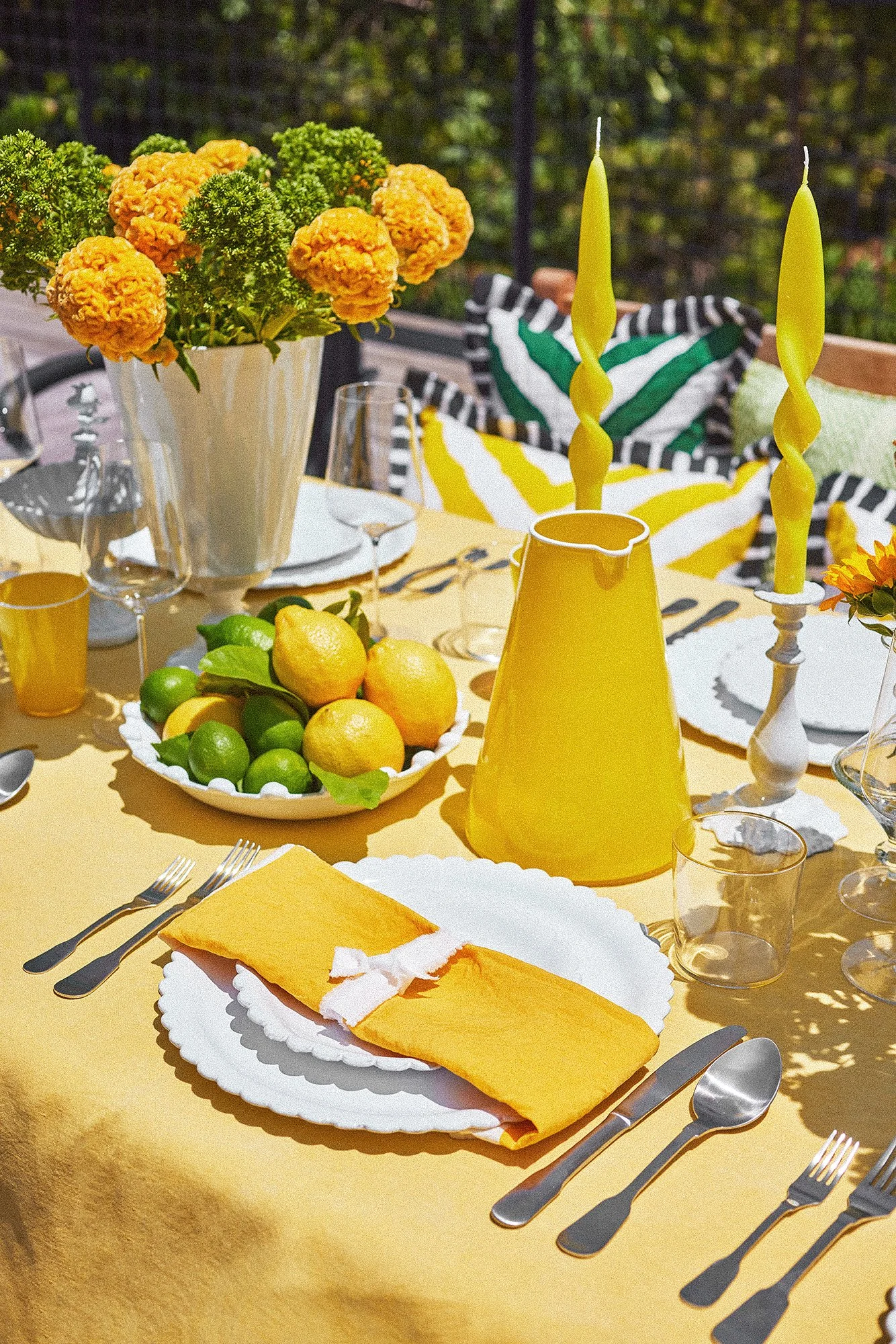 An outdoor dining table set with yellow tablecloth, white plates, silverware, and a yellow pitcher with twisted yellow candles. A bowl of lemons and limes, a large white vase with orange and green flowers, and colorful striped pillows in the background.