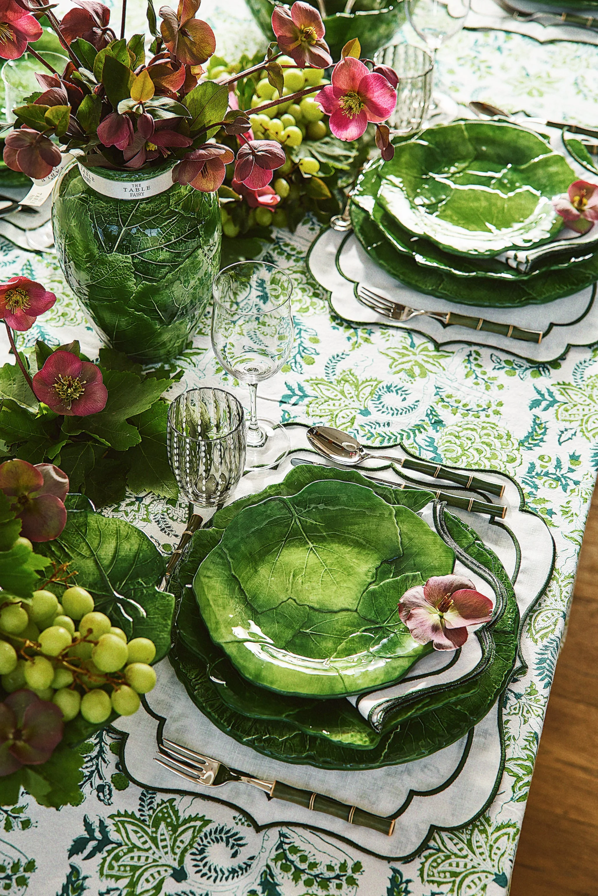 A table setting with green botanical-themed plates, glassware, and a floral centerpiece, featuring pink flowers and grape clusters on a patterned tablecloth.