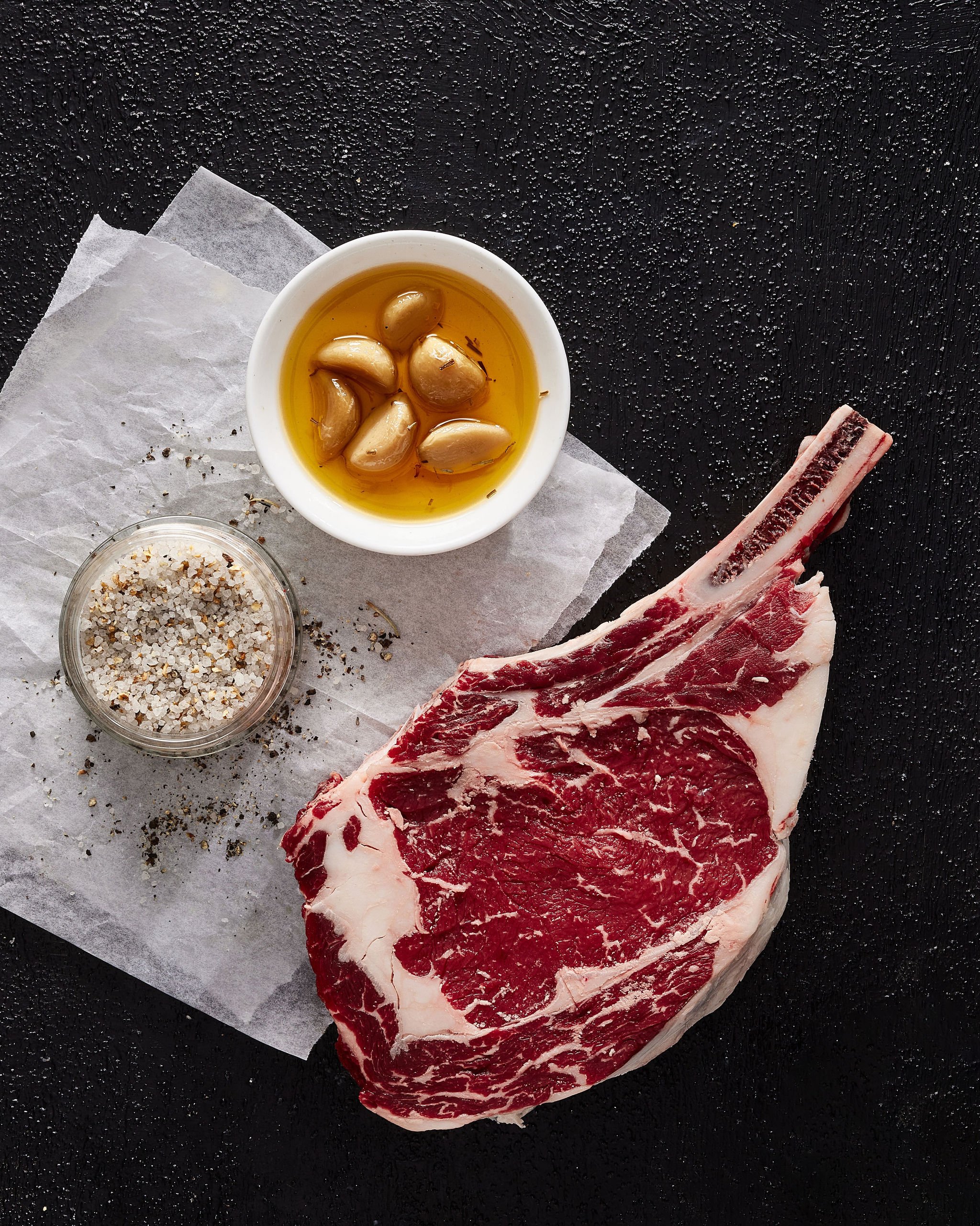 A raw beef ribeye steak with a large bone, a small jar of coarse salt, and a bowl of garlic cloves soaked in olive oil