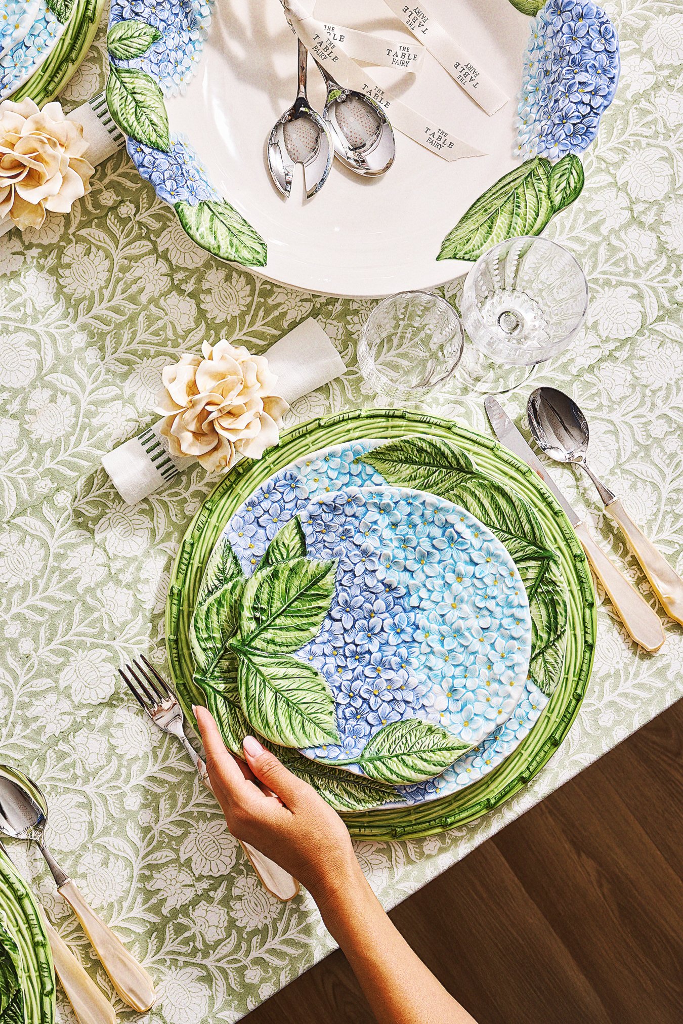 A table setting with a floral plate, matching napkin, and silverware, on a green and white patterned tablecloth. There are decorative leaves on the plates, and a person's hand holding a fork.