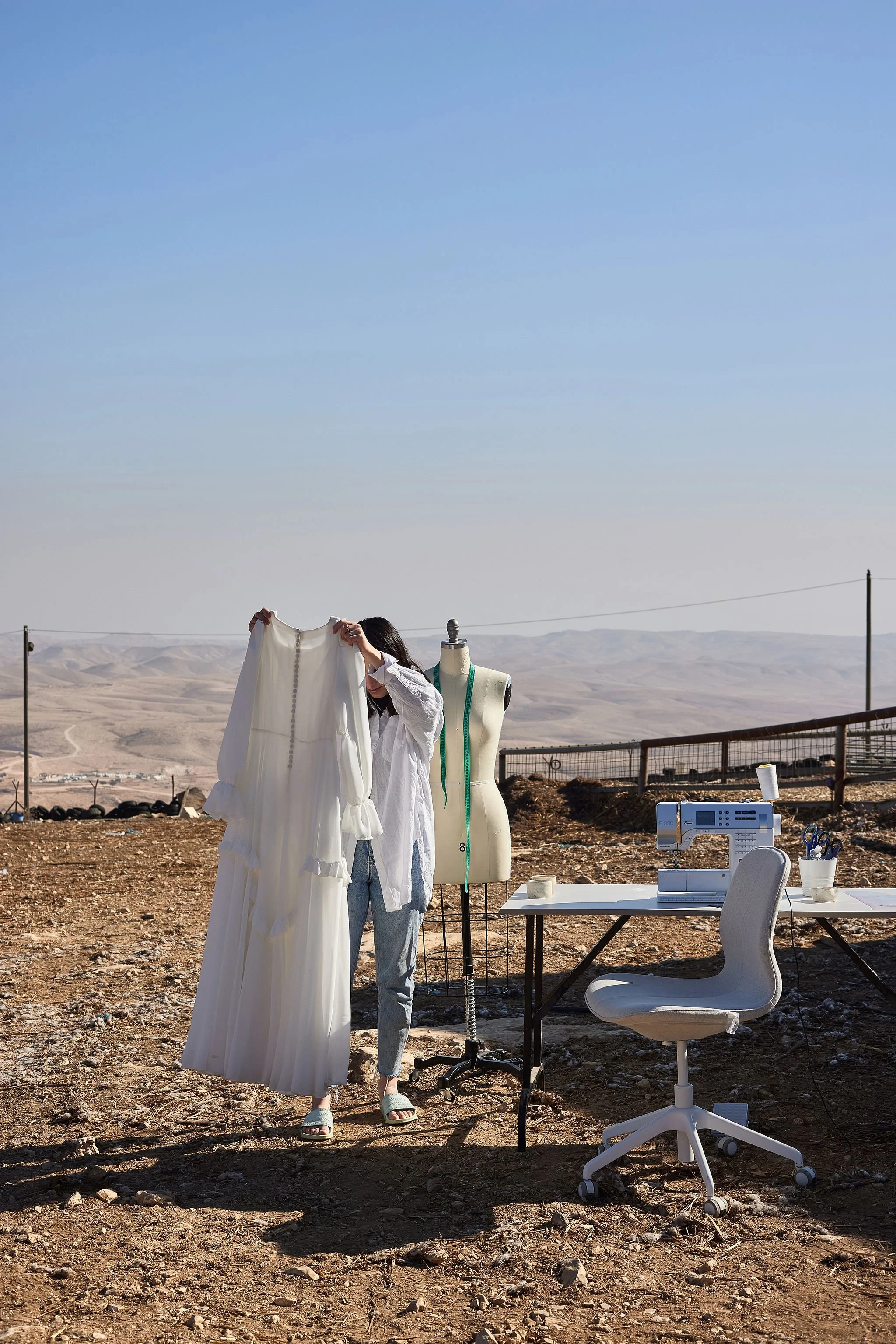 A person in a white shirt and jeans holding up a long white dress outdoors on a rocky, desert-like landscape with a mannequin, sewing machine, and office chair nearby.