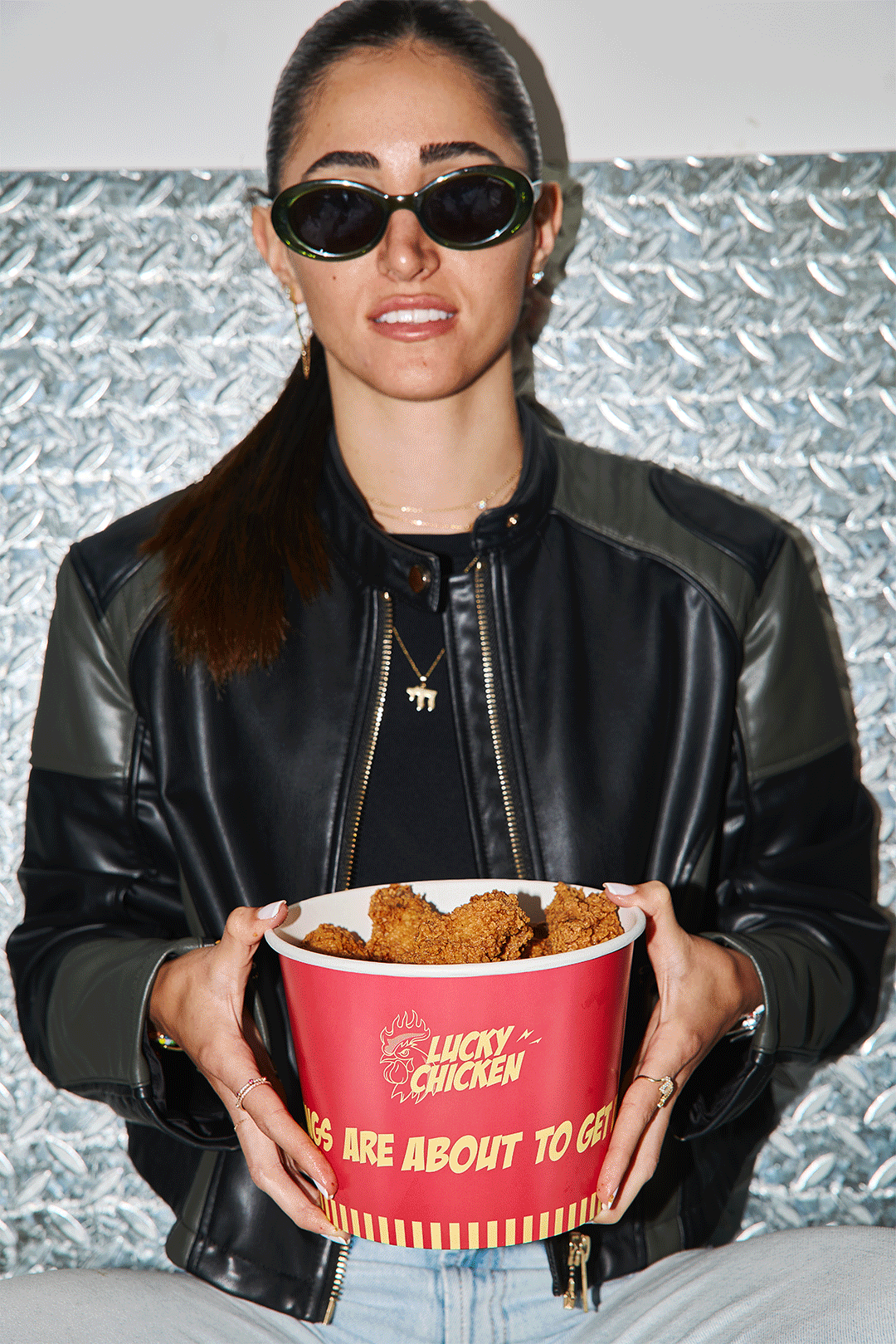 A woman wearing black sunglasses, a black leather jacket with green details, and a necklace with a dog pendant, holding a large red bucket of fried chicken from Lucky Chicken, in front of a metallic background.