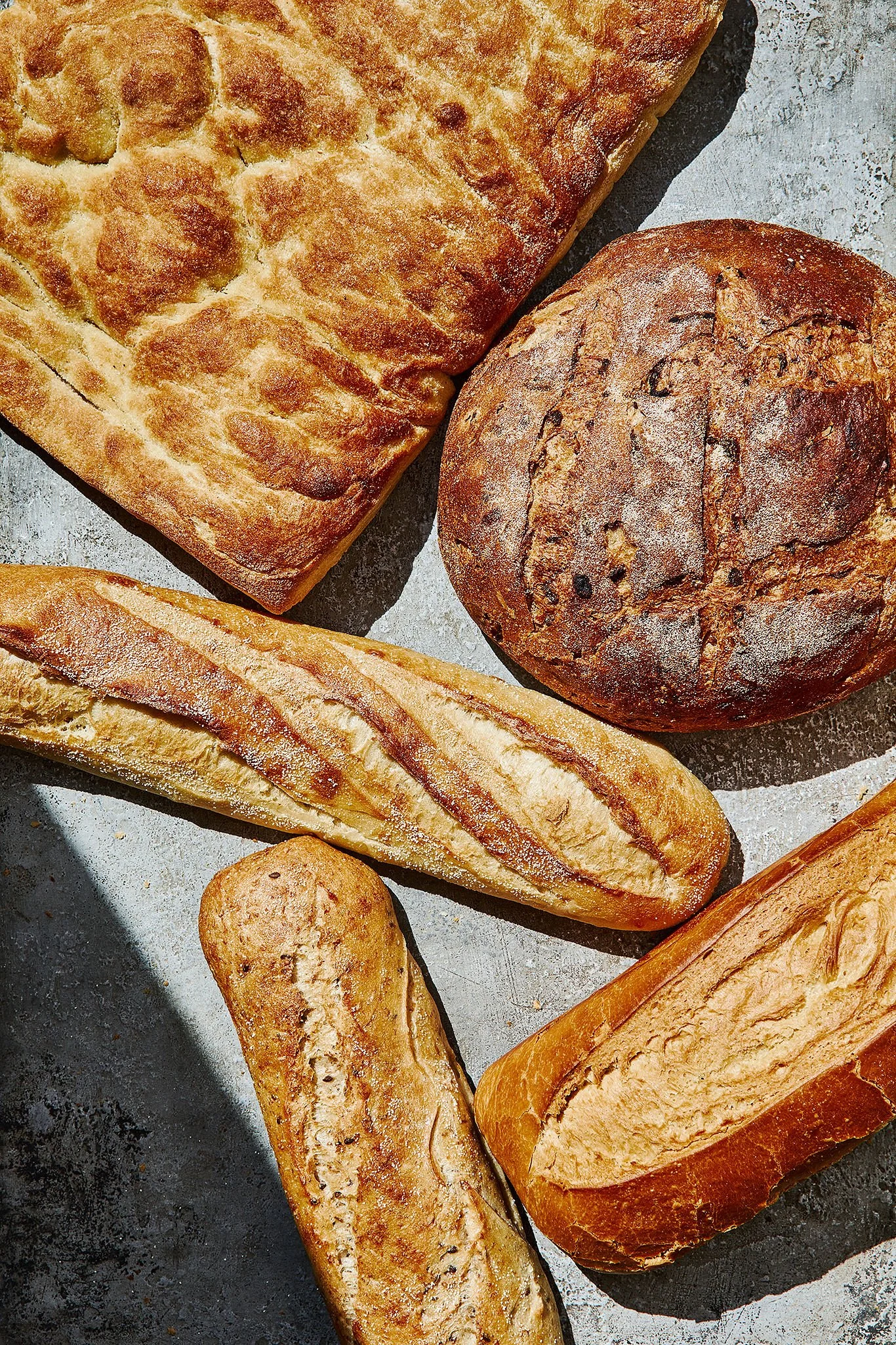 An assortment of freshly baked bread including a rectangular focaccia, round sourdough, baguette, sandwich bread, ciabatta, and baguette on a textured surface.