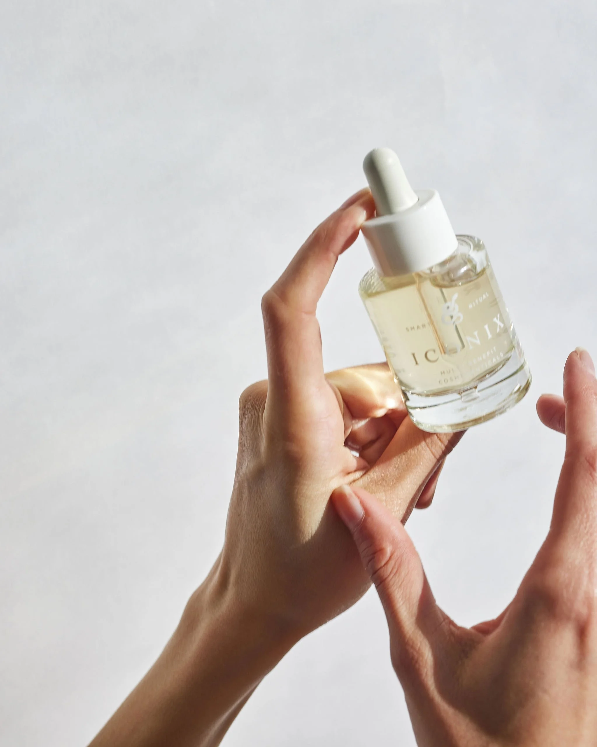 A person's hand holding a clear glass dropper bottle with light-colored liquid, labeled 'ICONIX' against a plain white background.