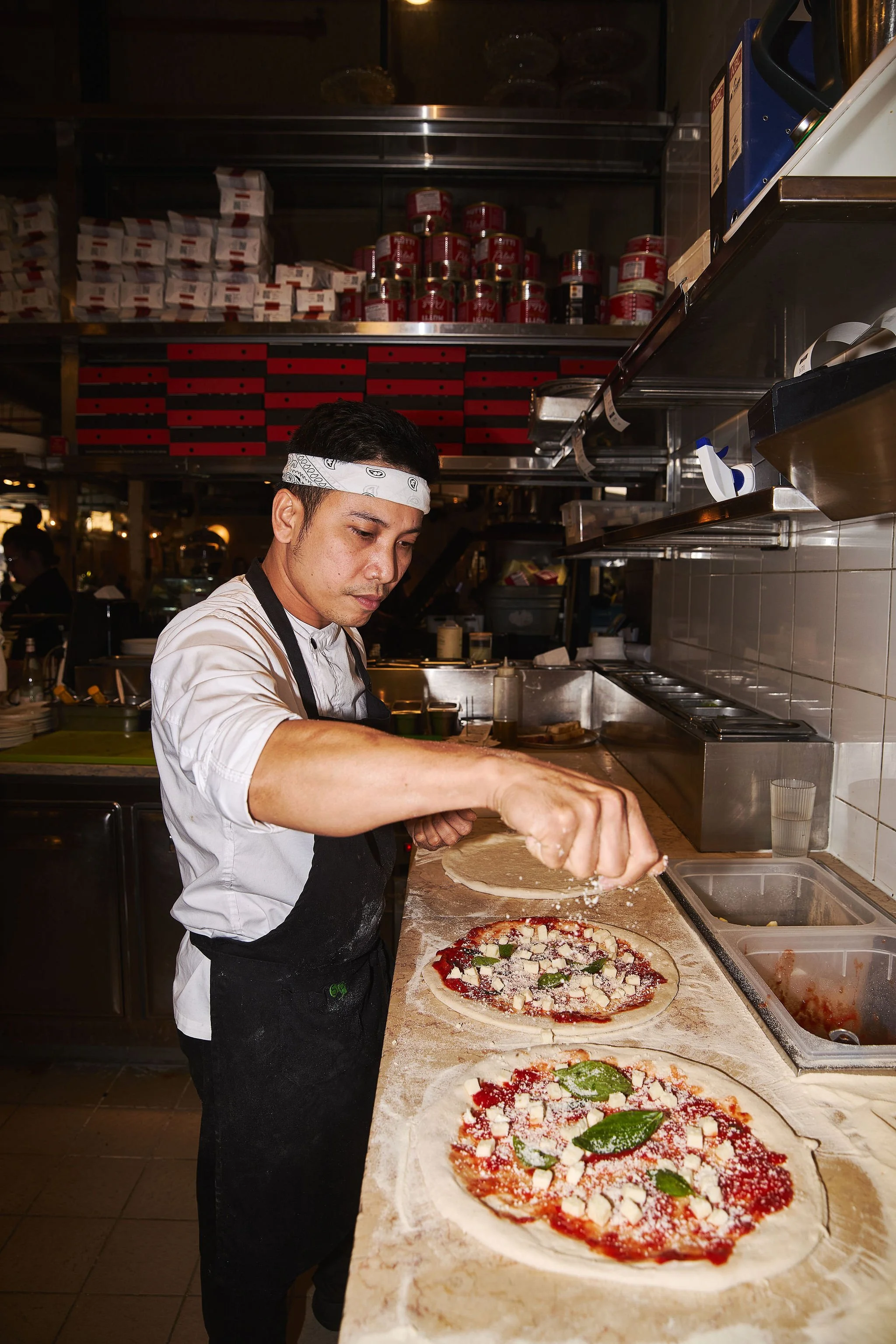 A chef prepares pizza in a restaurant kitchen, sprinkling cheese or seasoning onto two uncooked pizzas topped with tomato sauce, cheese, basil leaves, and other ingredients.