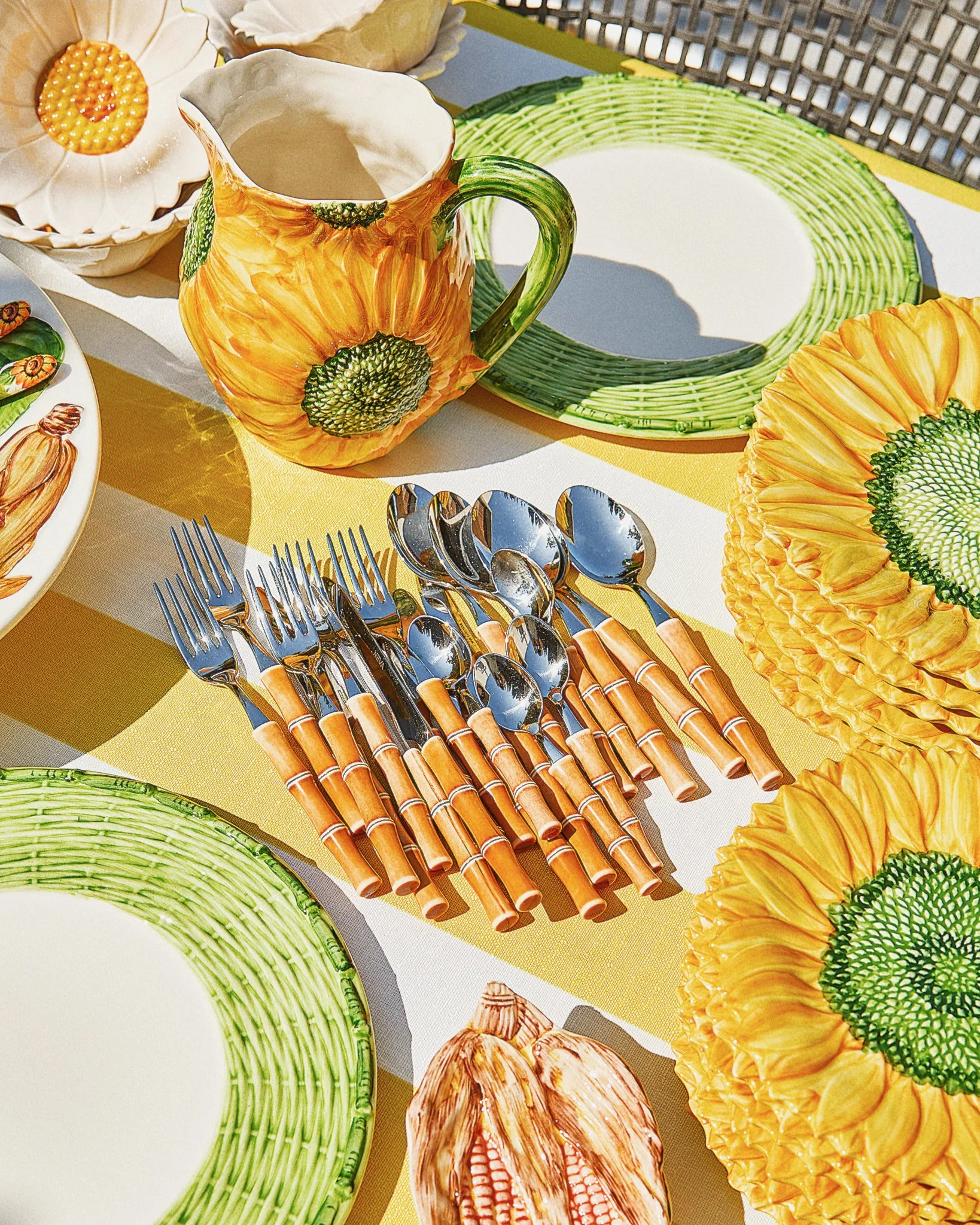 Decorative sunflower-themed tableware including a sunflower-patterned pitcher, large sunflower-patterned plates, zebra-striped and green-bordered dishes, and an assortment of silverware with bamboo-style handles arranged on a yellow and white striped