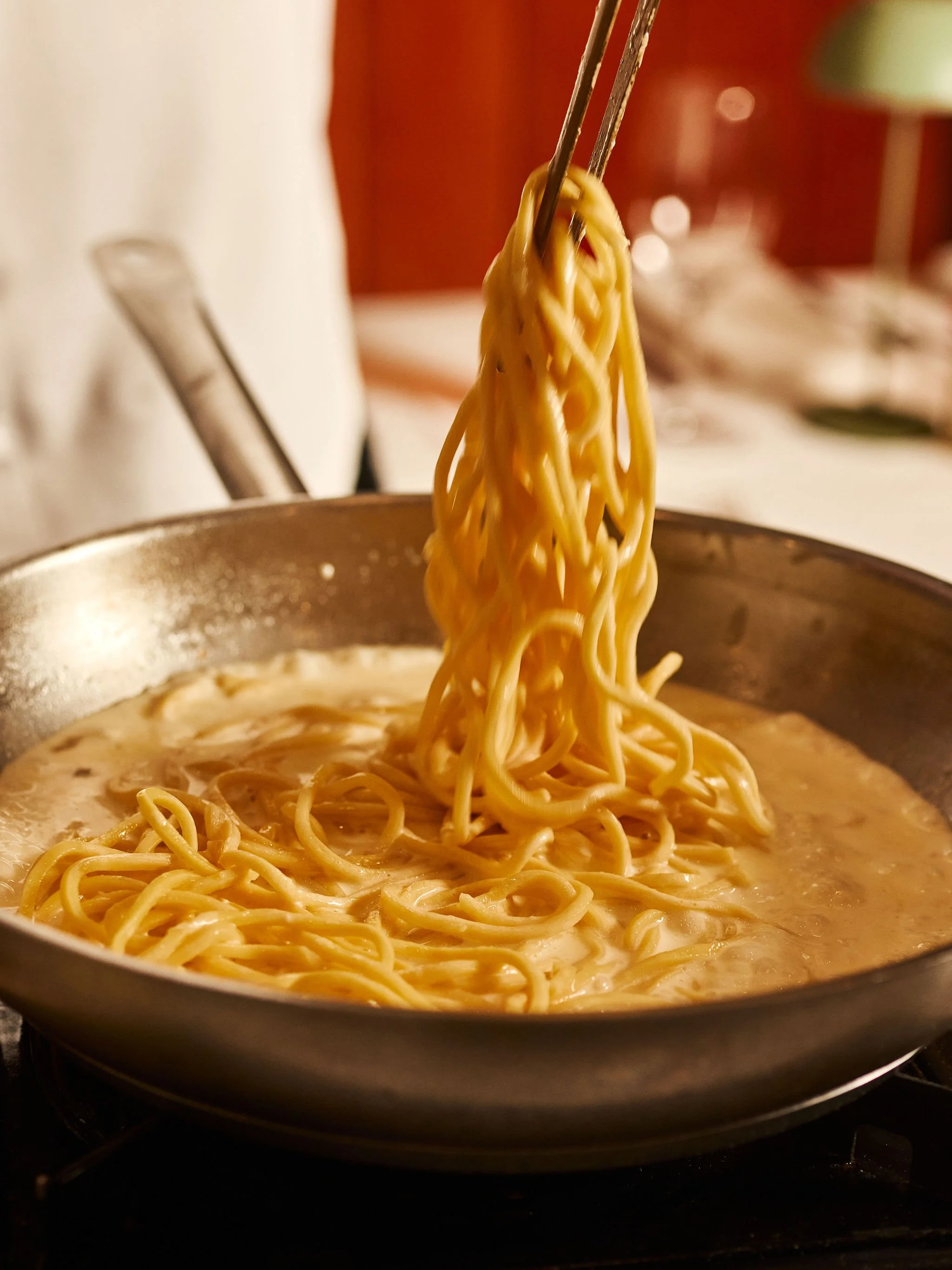 A close-up of spaghetti noodles being lifted by tongs from a pan with creamy sauce.