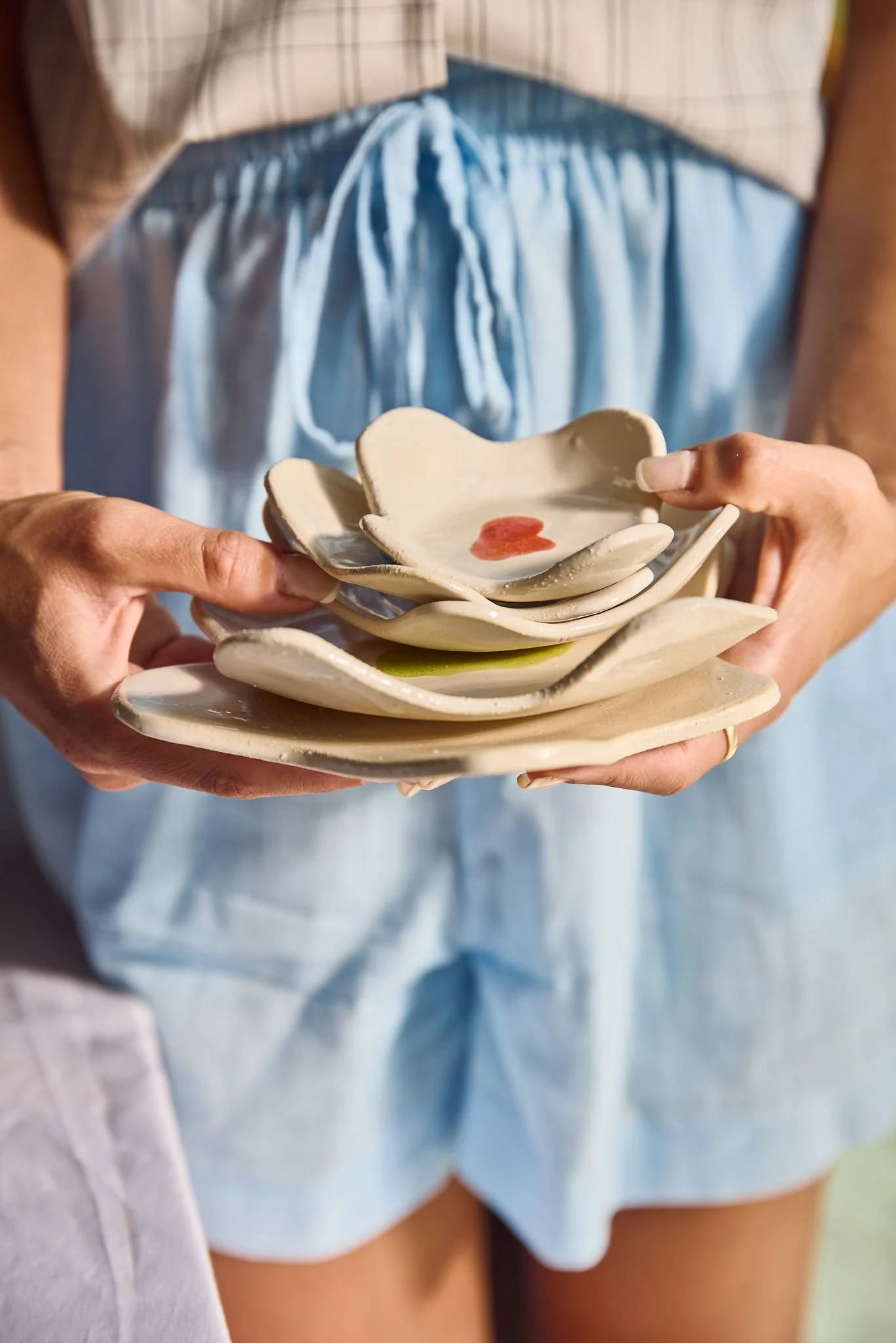 Person holding stacked ceramic bowls with a heart design, standing in front of a person wearing light blue shorts and a beige striped shirt.