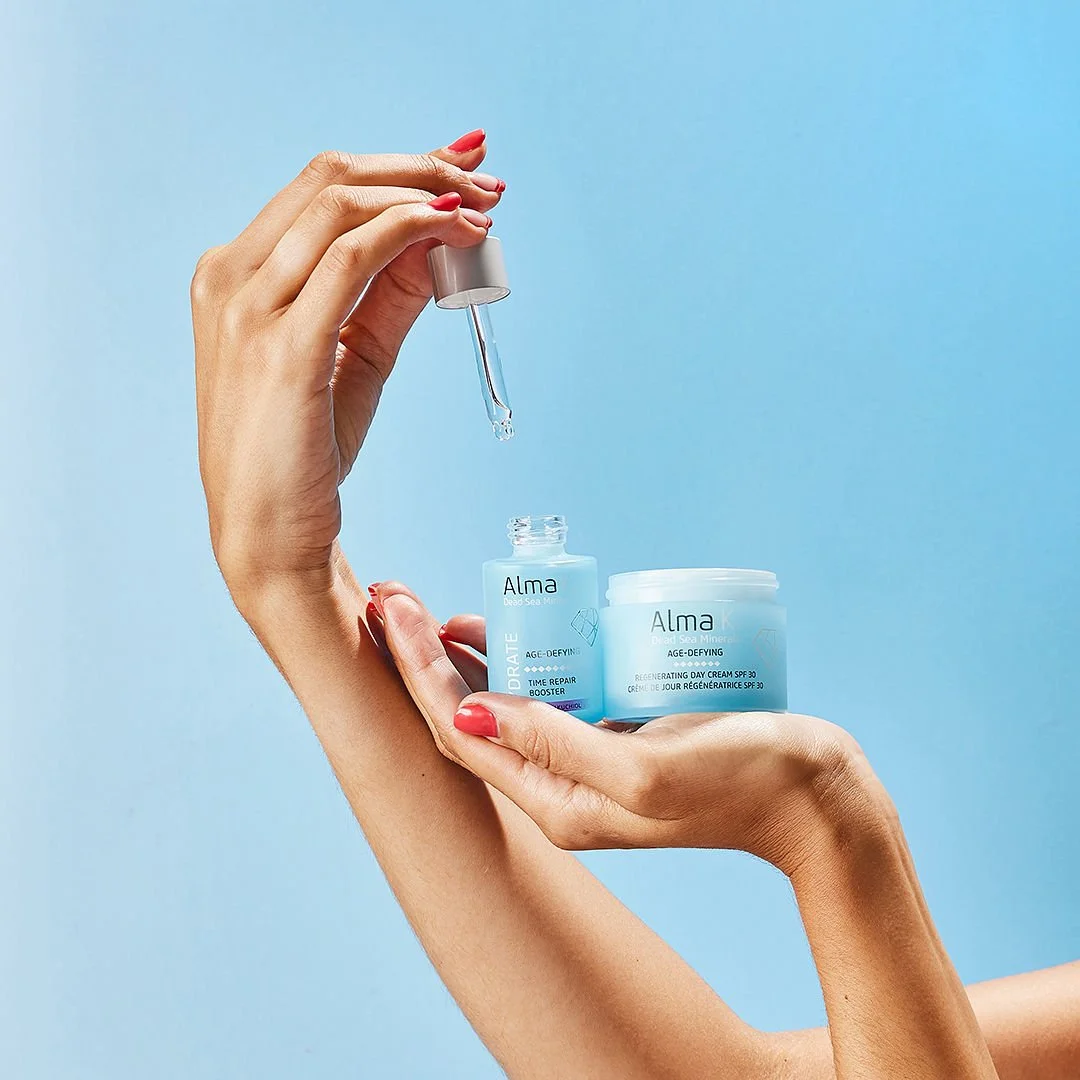 Hands holding skincare products, one with a dropper dispensing serum into another container labeled 'Alma,' with a light blue background.