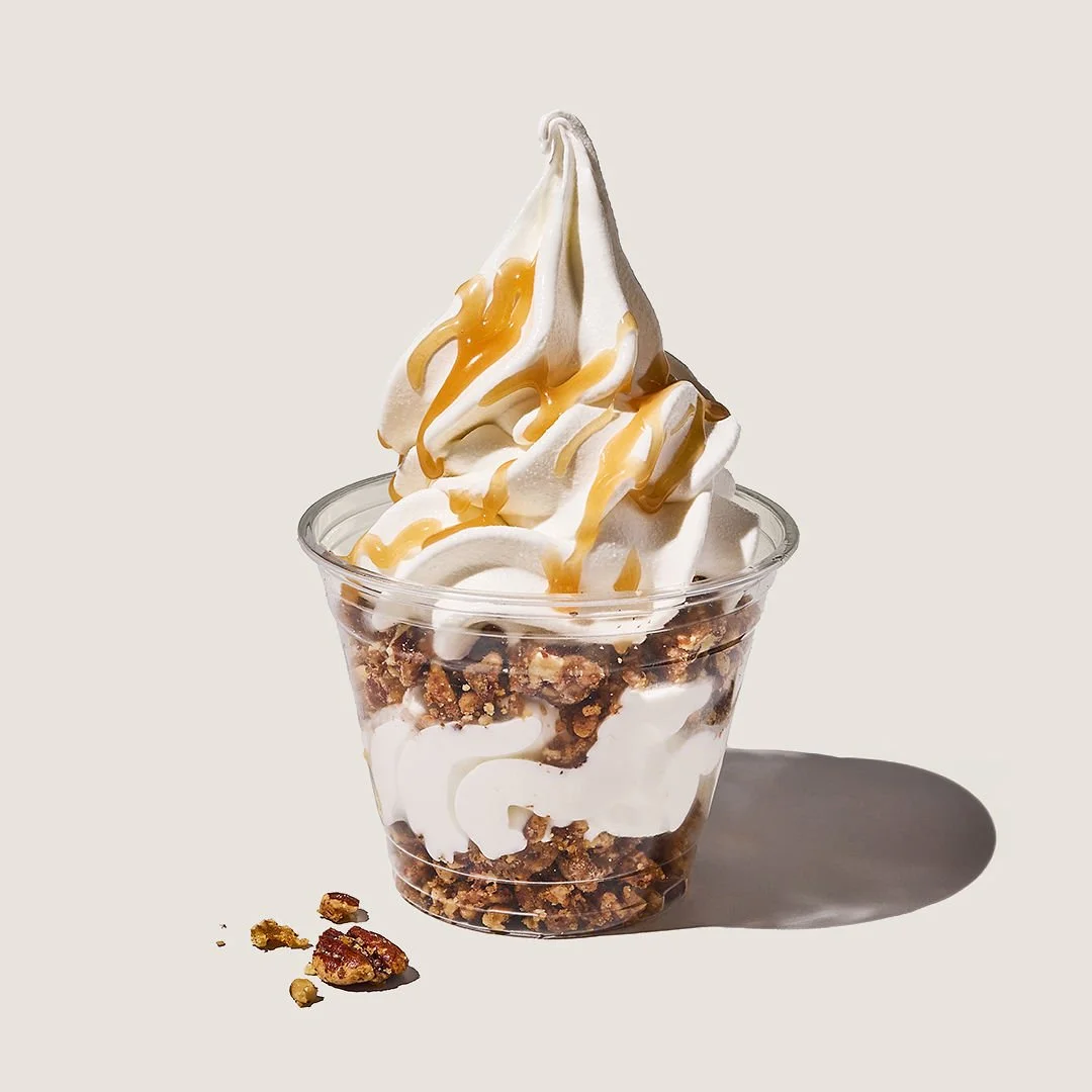 Soft serve ice cream with caramel drizzle in a clear cup, layered with granola and caramel, placed on a white surface with a shadow.