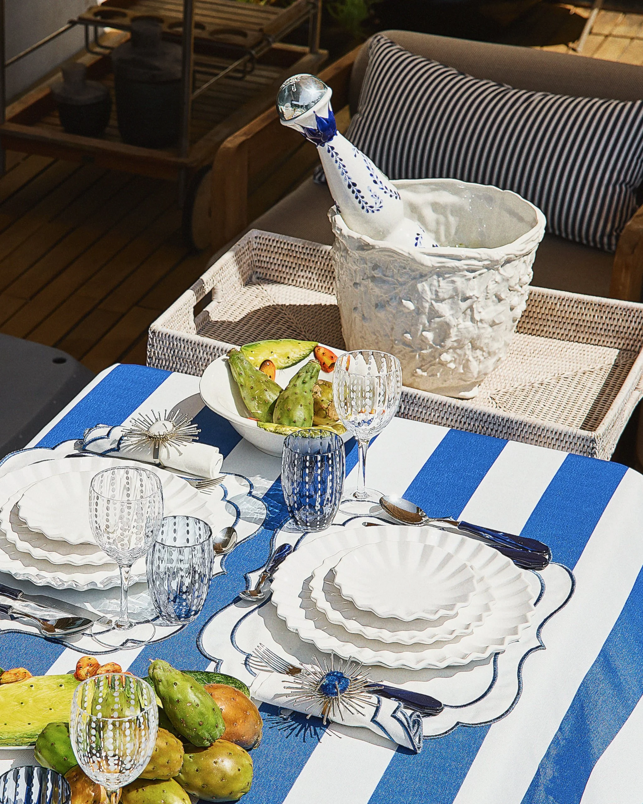 Outdoor dining table set with white and blue tableware, glassware, and a bowl of prickly pear fruit. In the background, a decorative ice bucket with a bottle inside is on a wicker tray, with a sofa and striped pillow beside it.